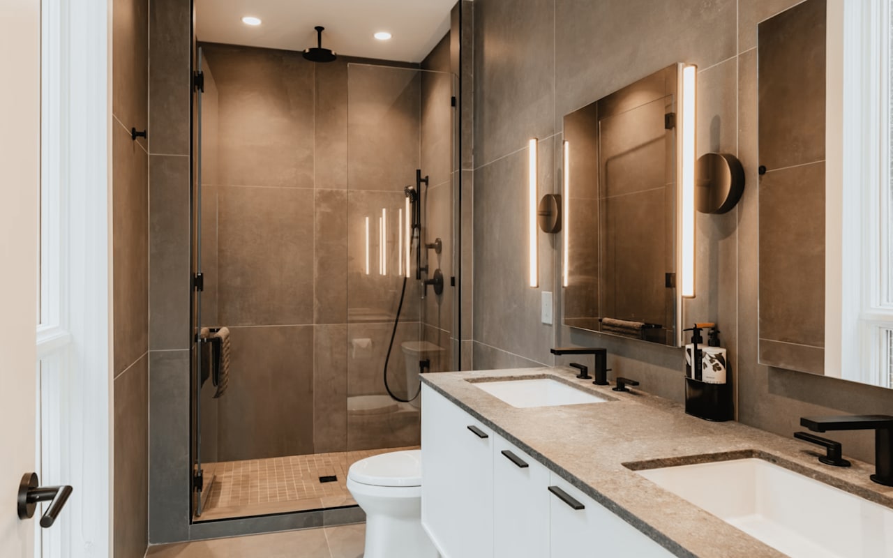 Modern bathroom with double sink vanity, illuminated mirrors, and a glass-enclosed shower with black fixtures.