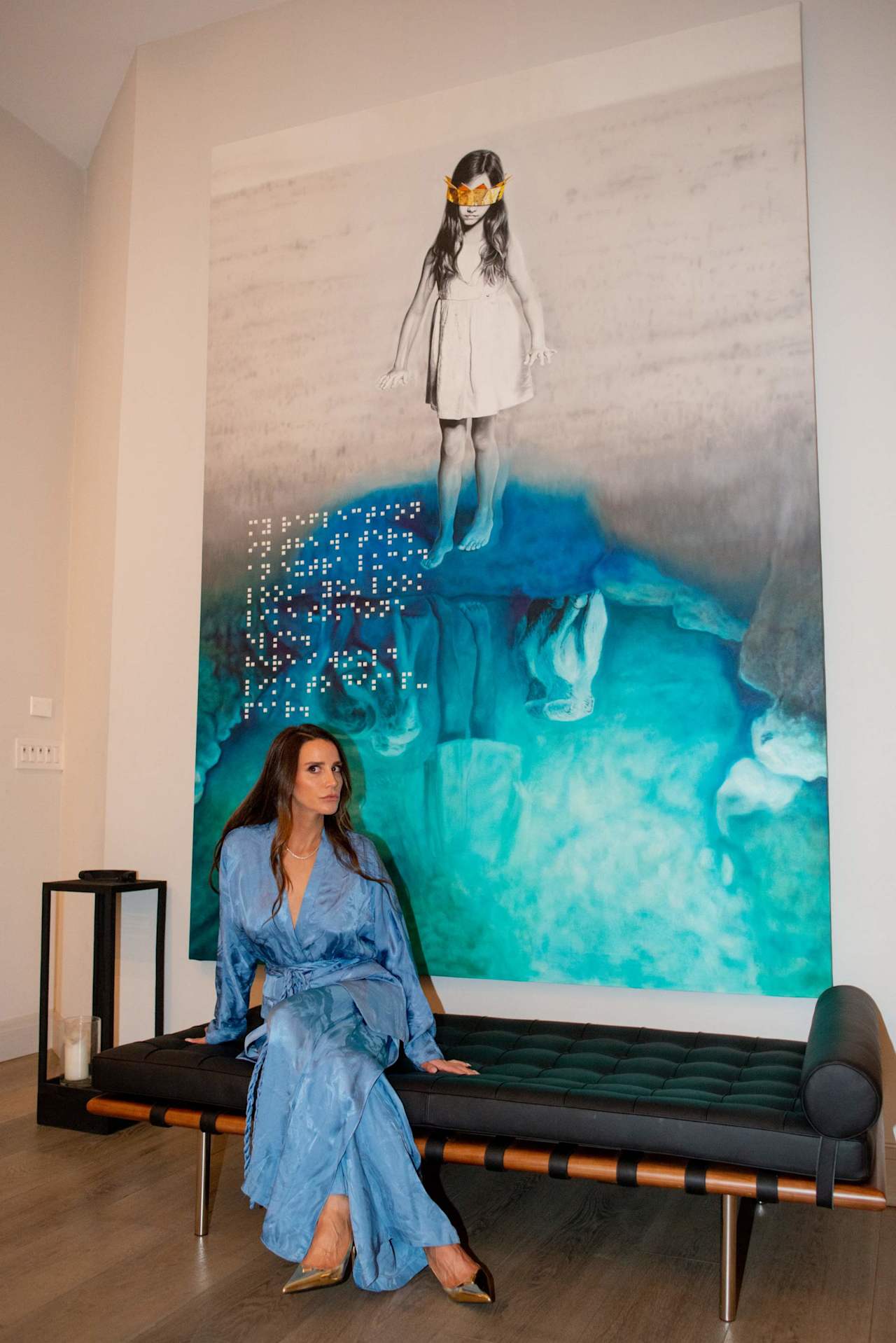Woman in a blue dress seated on a black sofa beneath a large colorful artwork.