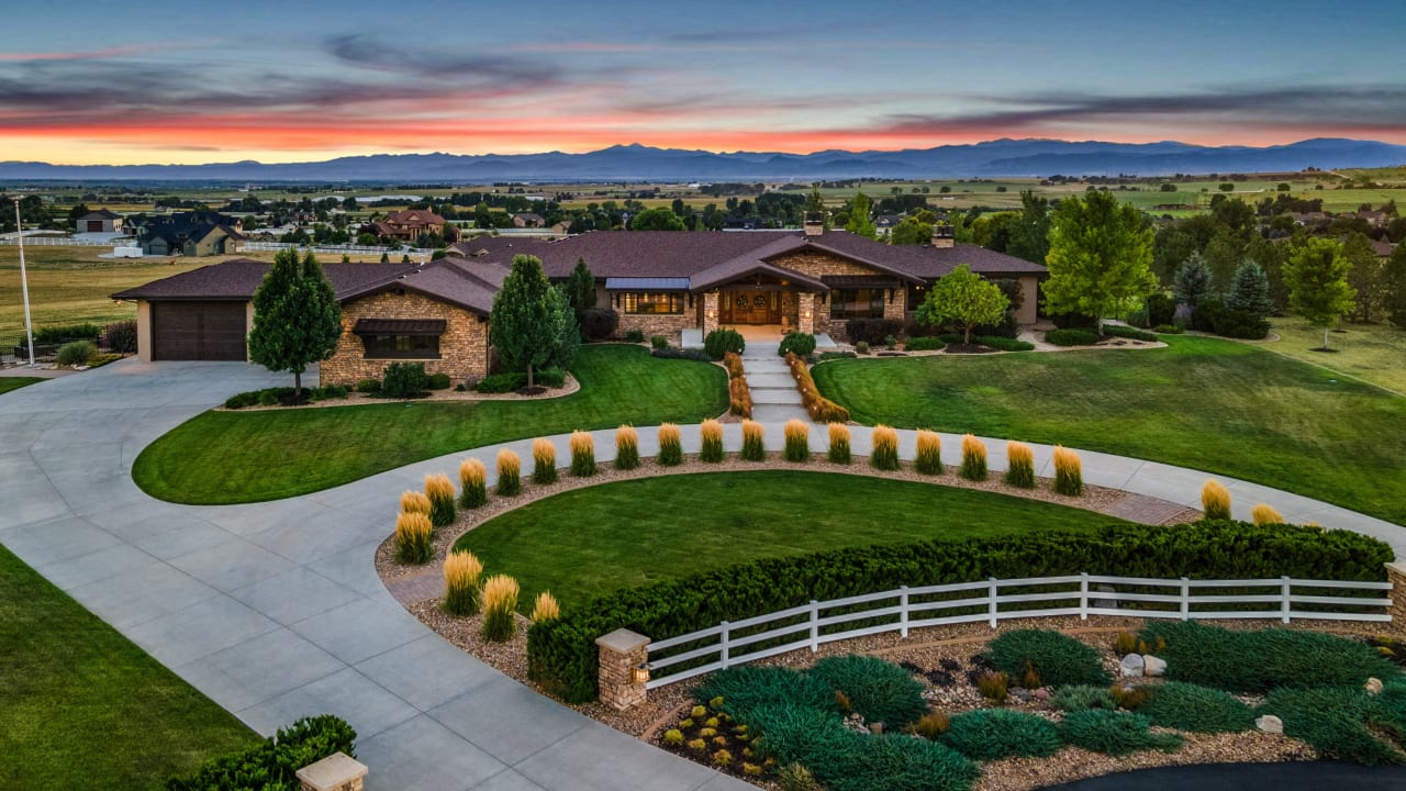 The facade of a stone and wood prairie-style home is photographed with impeccable, modern landscaping. The sunset is visible in the background with Colorado mountain views.