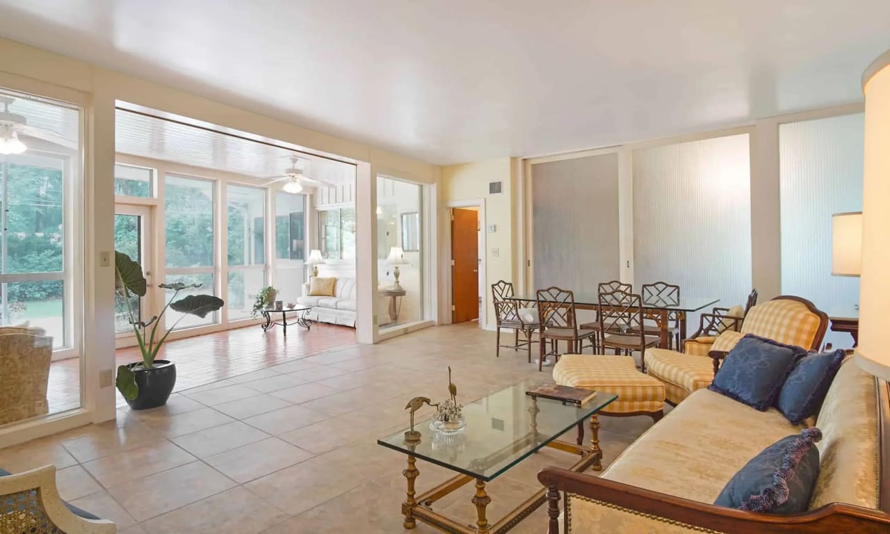 An interior view of a living space with large windows, a tile floor, and comfortable seating. The room is well-lit and has a few decorative plants.