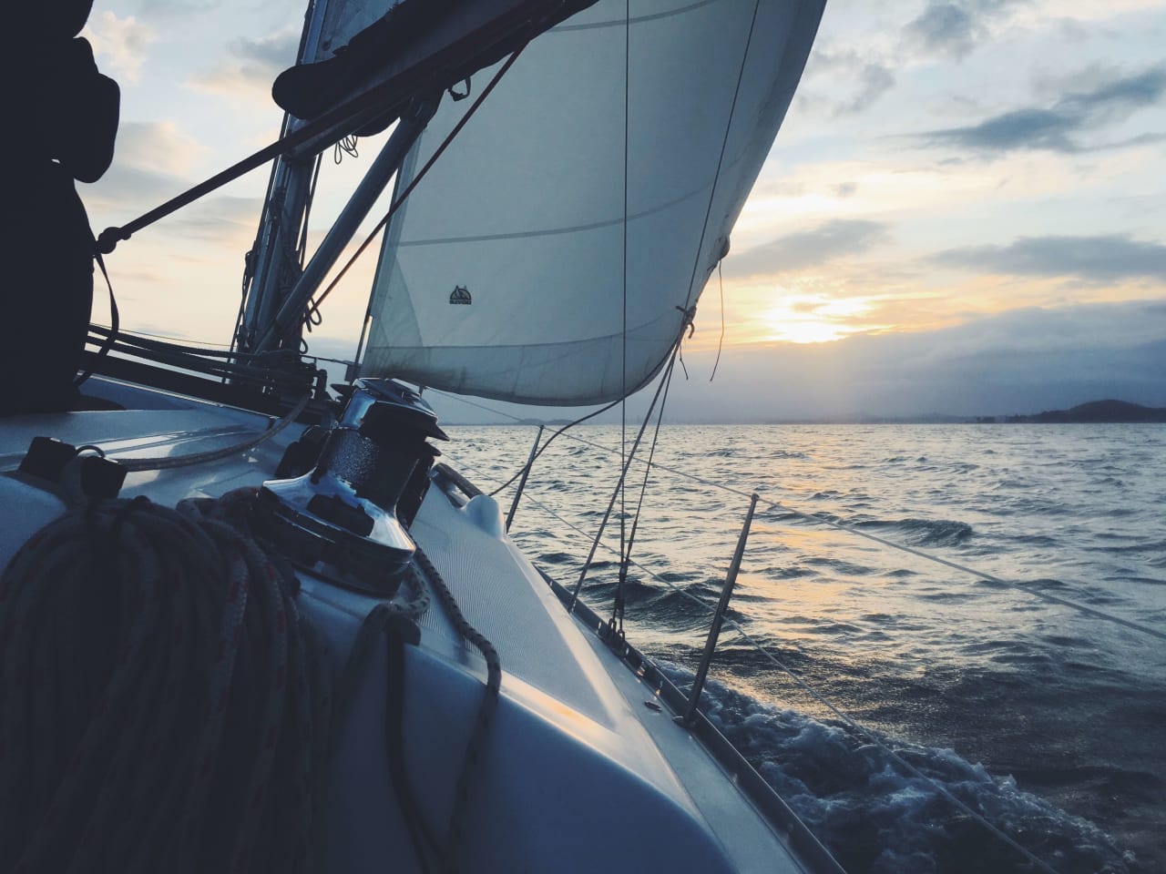 A sailboat with white sails slicing through the calm ocean waters at sunset.