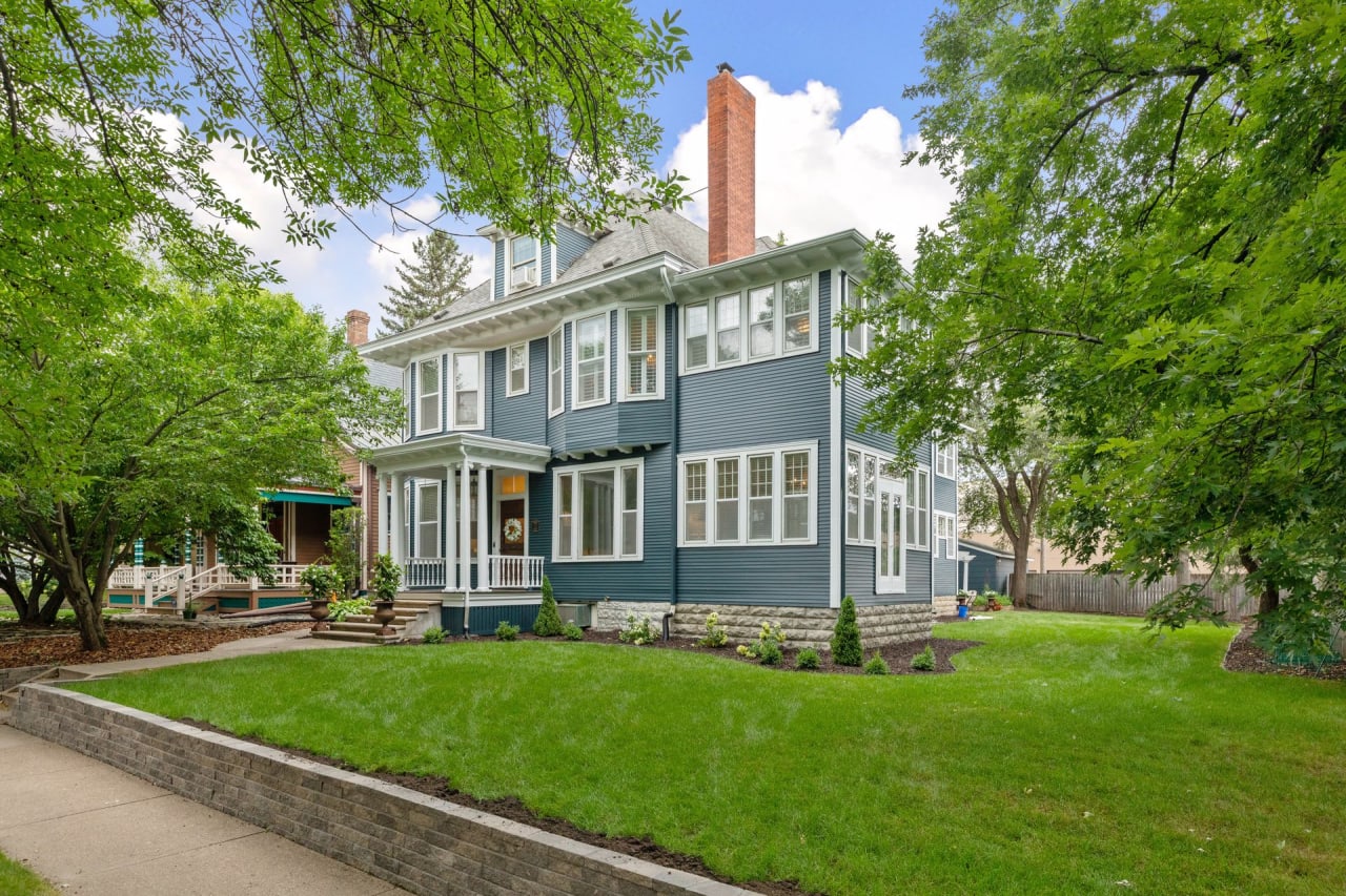 A blue house with a chimney, a lush green yard with plants, and large trees.