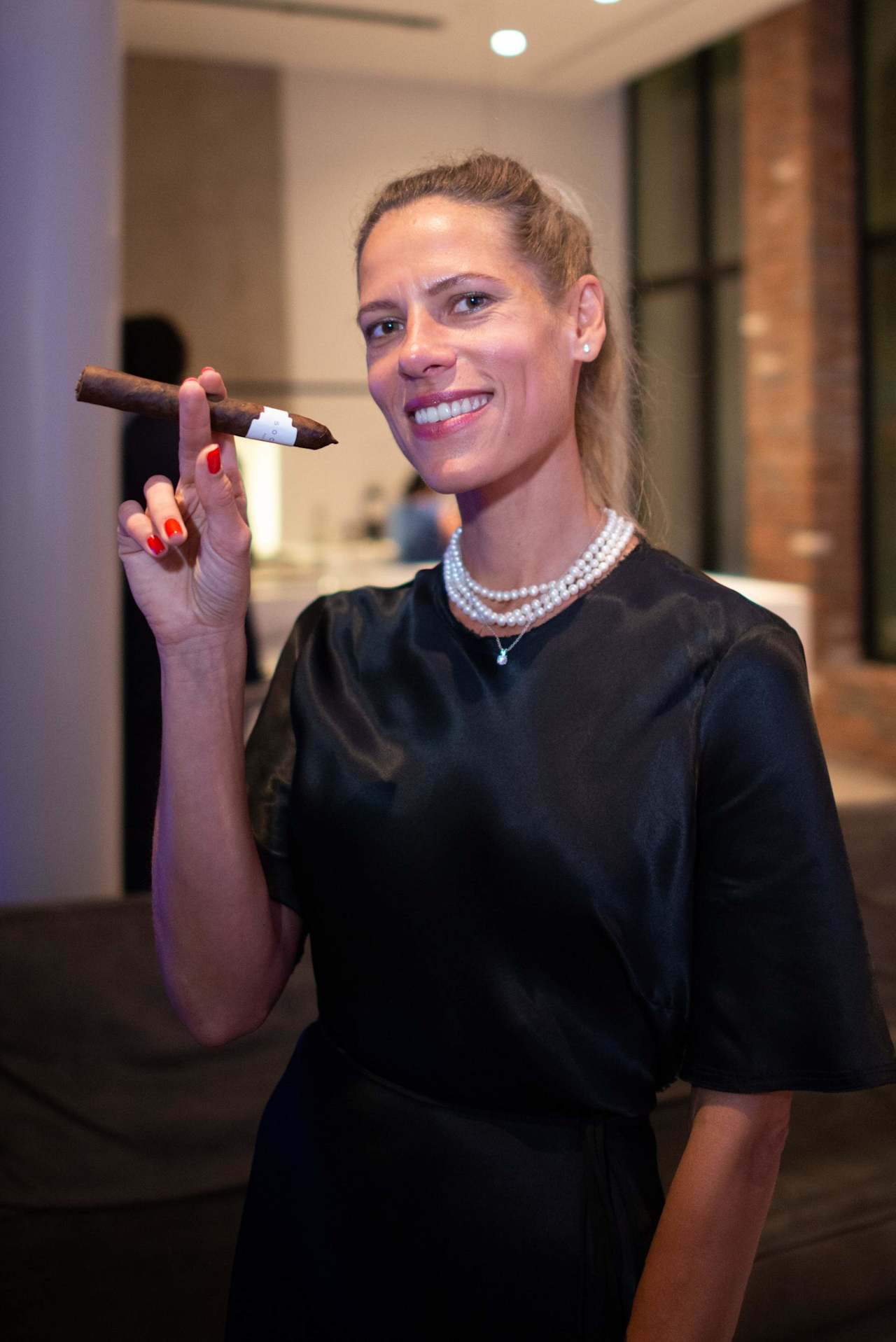 Smiling woman in a black dress and layered pearls holding a cigar at an indoor event.