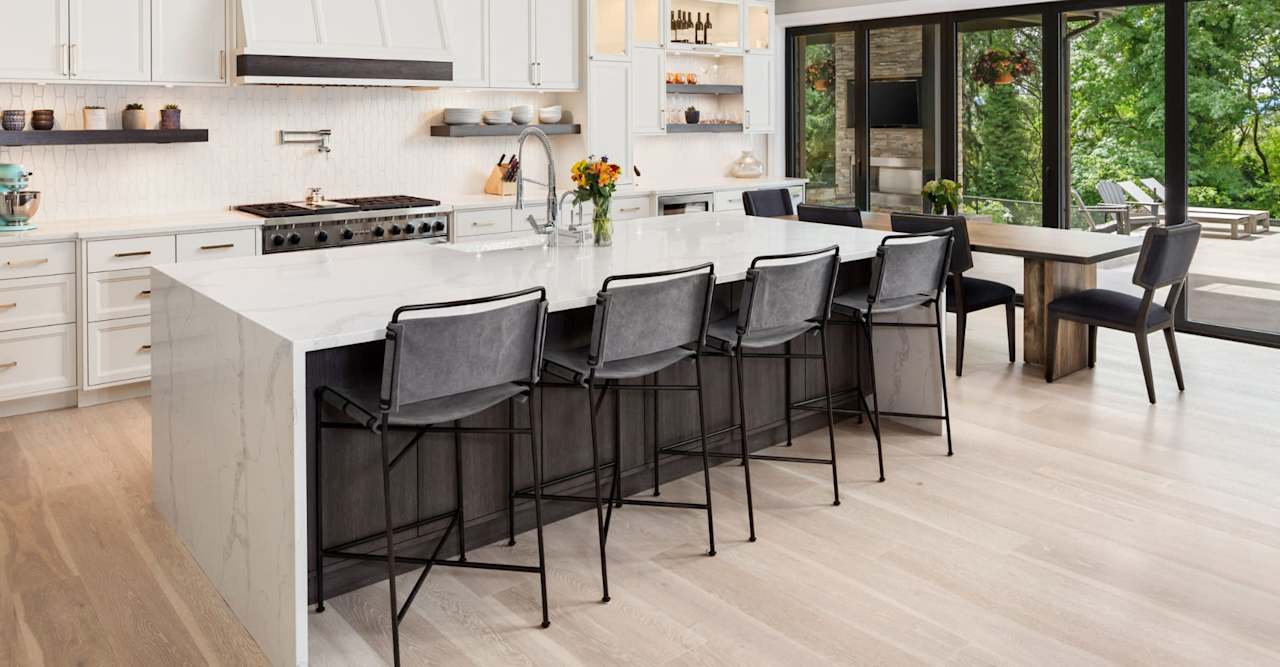 Modern white kitchen with a large island, dark barstools, and glass doors opening to a wooded patio.