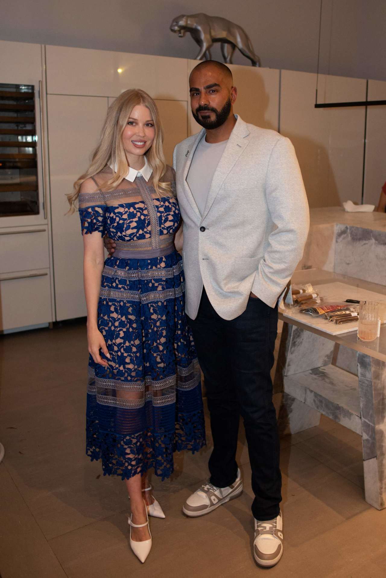 A woman in a blue lace dress and a man in a light blazer pose together indoors.