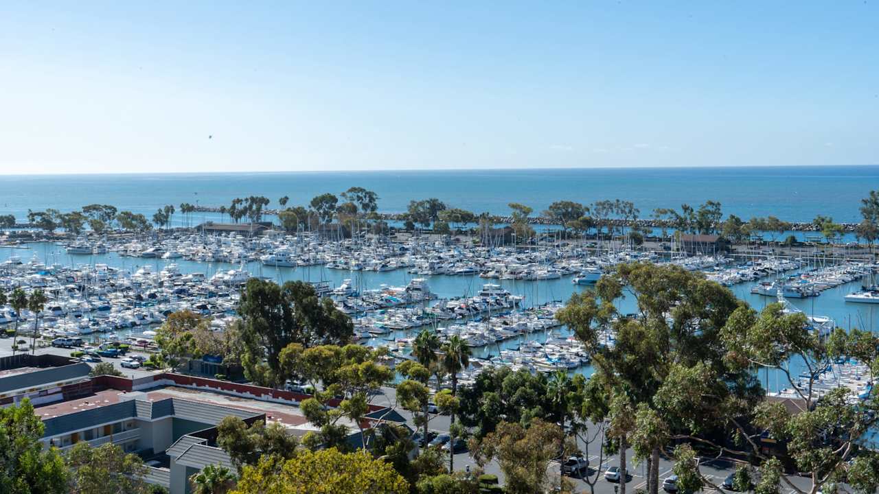 An aerial view of Dana Point Marina with boats of various sizes docked in the harbor and surrounded by a few buildings.