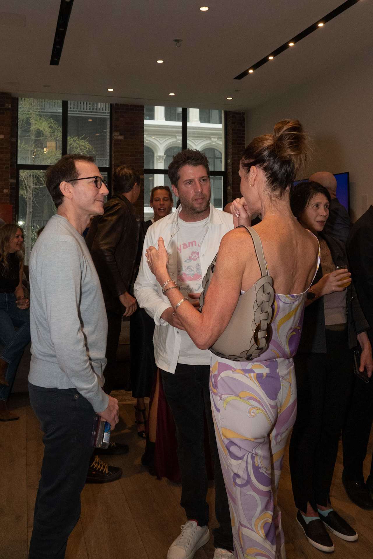 Adults mingle at an indoor gathering; a woman in a colorful patterned dress talks with two men.