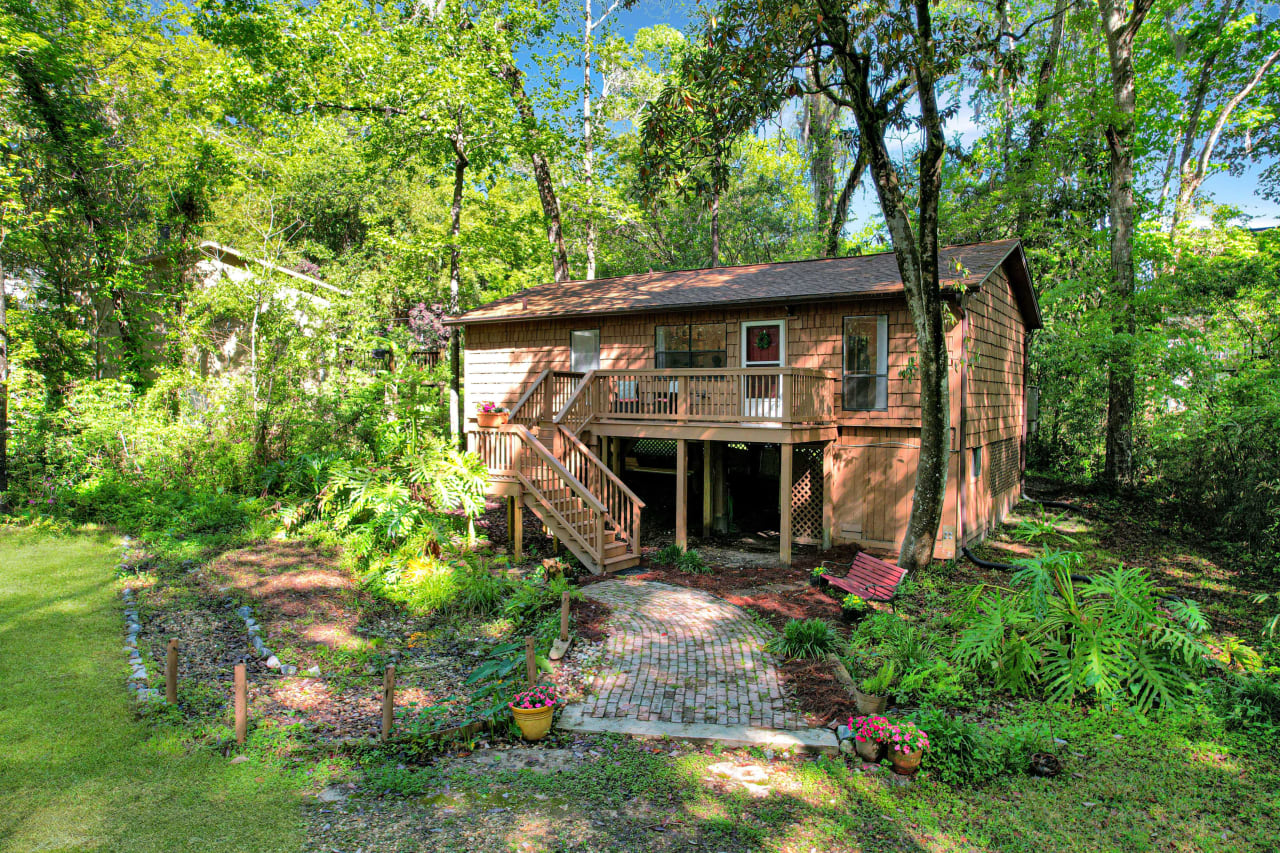 The exterior of a house surrounded by lush greenery. The house has a combination of brick and siding and is elevated with steps leading up to the entrance. The yard is well-maintained with a grassy lawn and some landscaping, creating a serene and private setting.