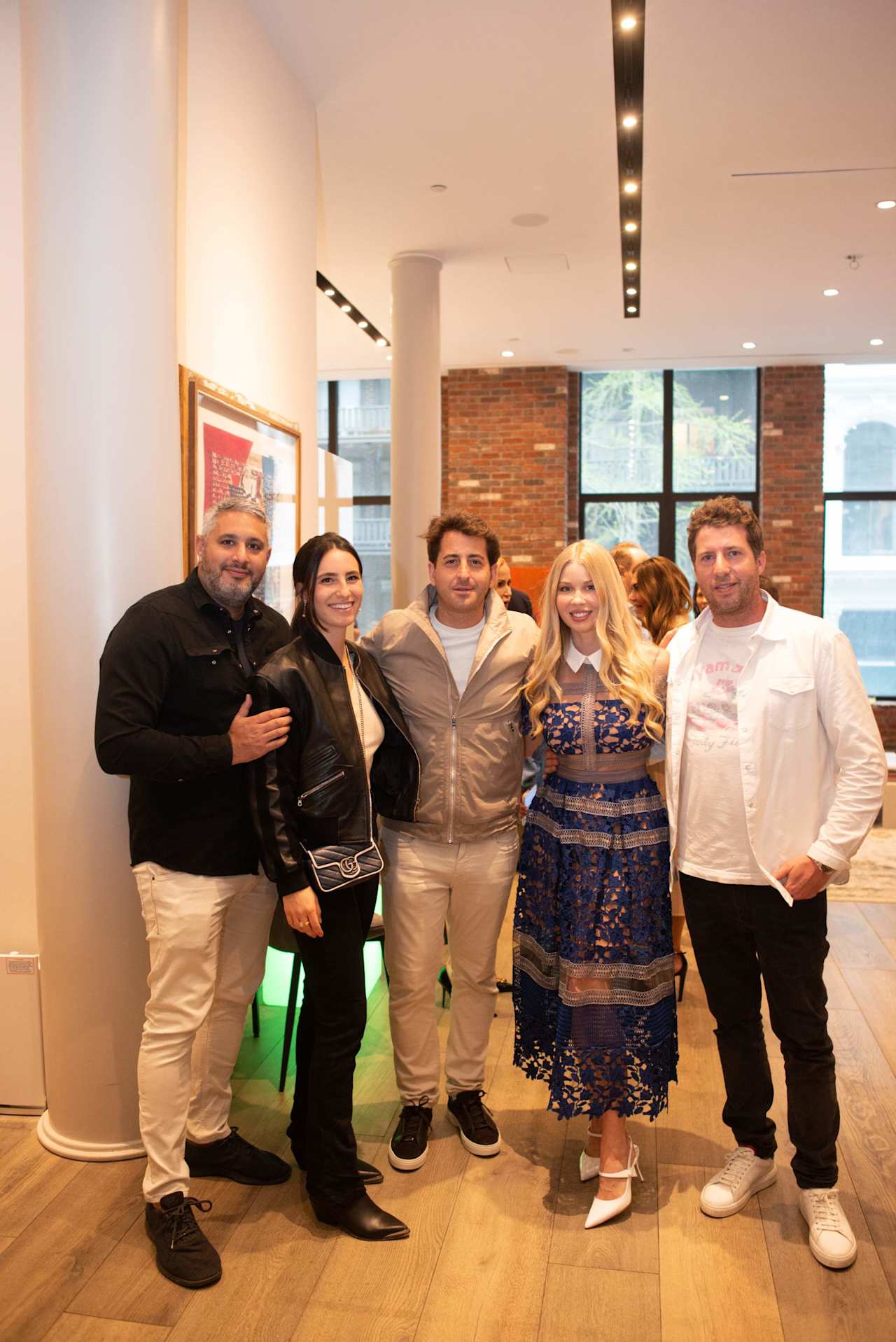 Five adults pose together at an indoor gathering, with brick walls and large windows behind them.