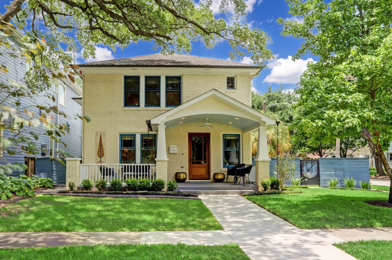 A charming, sunny two-story home with a welcoming porch nestled among mature trees.