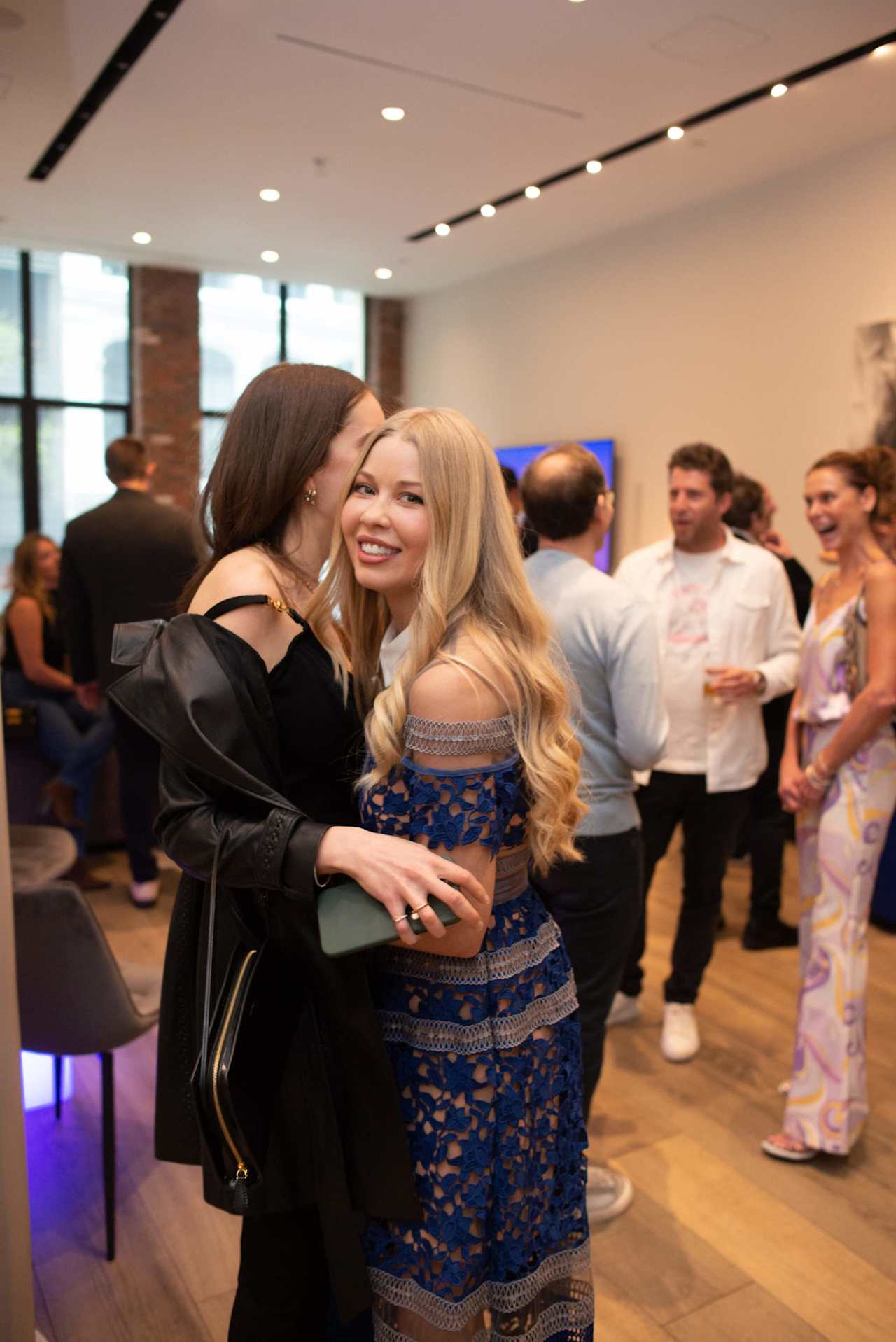 A woman in black embraces a smiling woman in a blue lace dress at a reception.