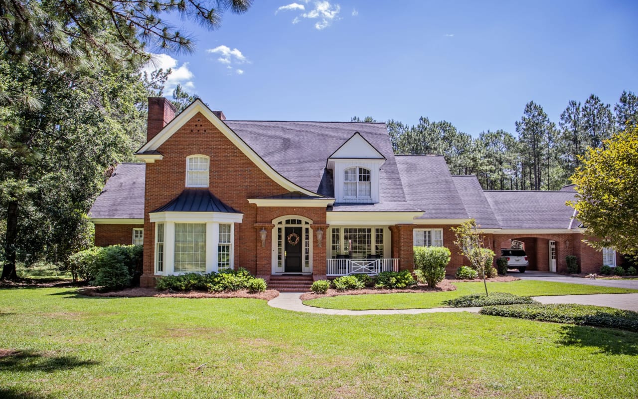 A large brick house with a white picket fence, paved walkway, two-car garage, front yard, and trees lining the street.