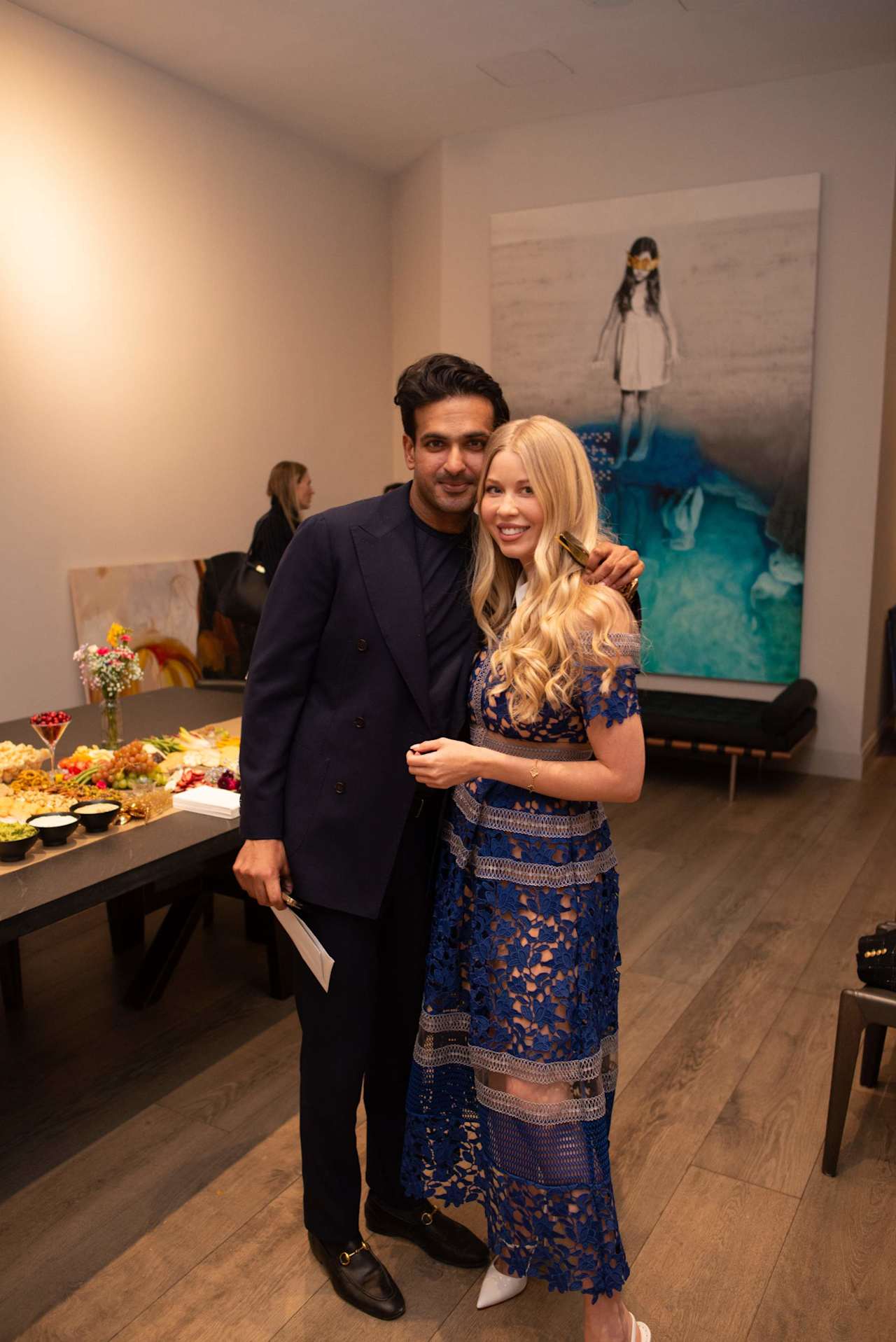 A man and woman pose together at an event, with a buffet and large artwork behind them.