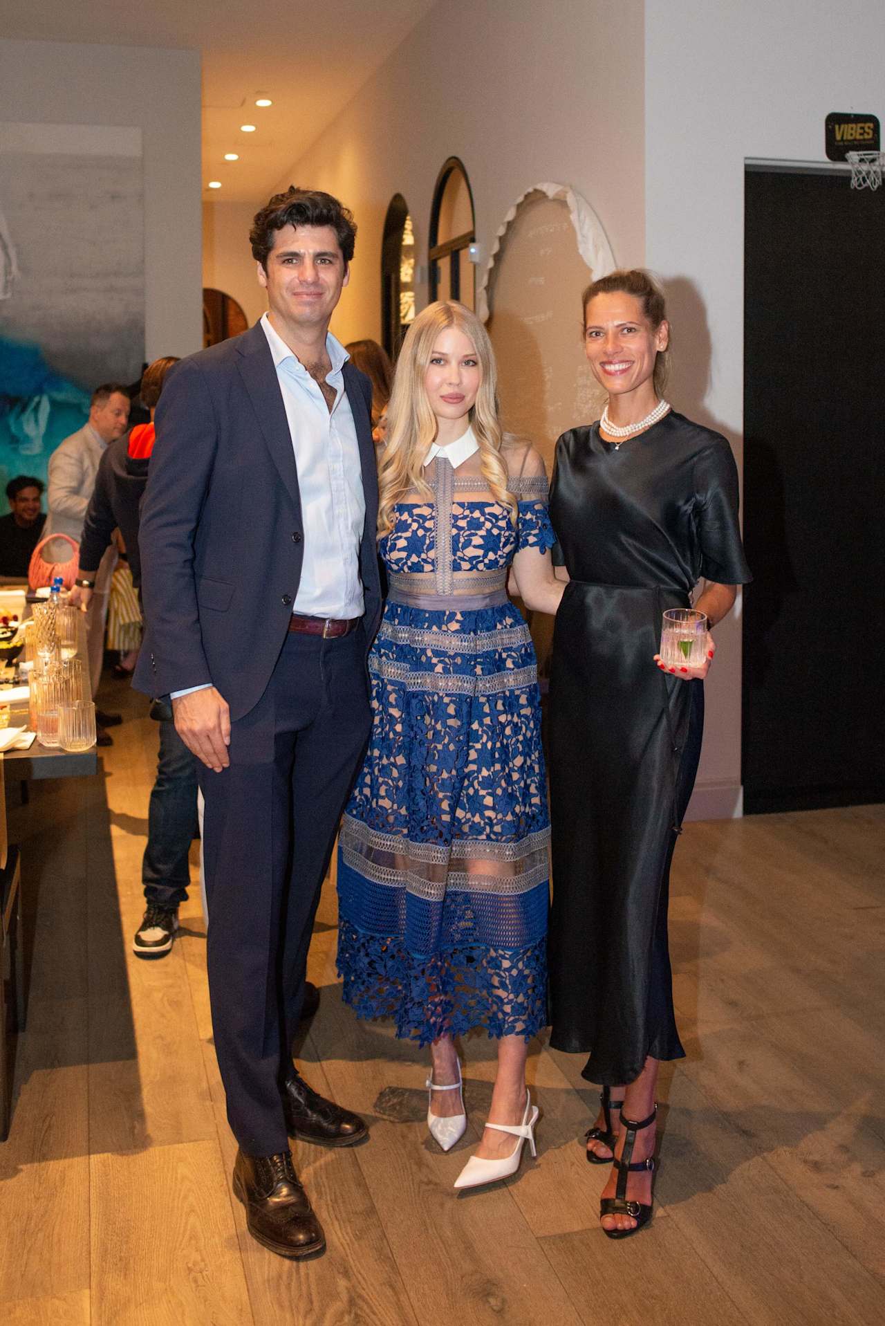 Three adults pose at an indoor event; the center woman wears a blue lace dress.