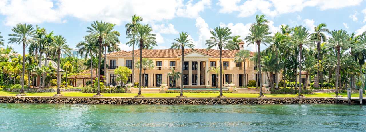 Waterfront Mediterranean-style home framed by palms, with landscaped grounds and a stone seawall.