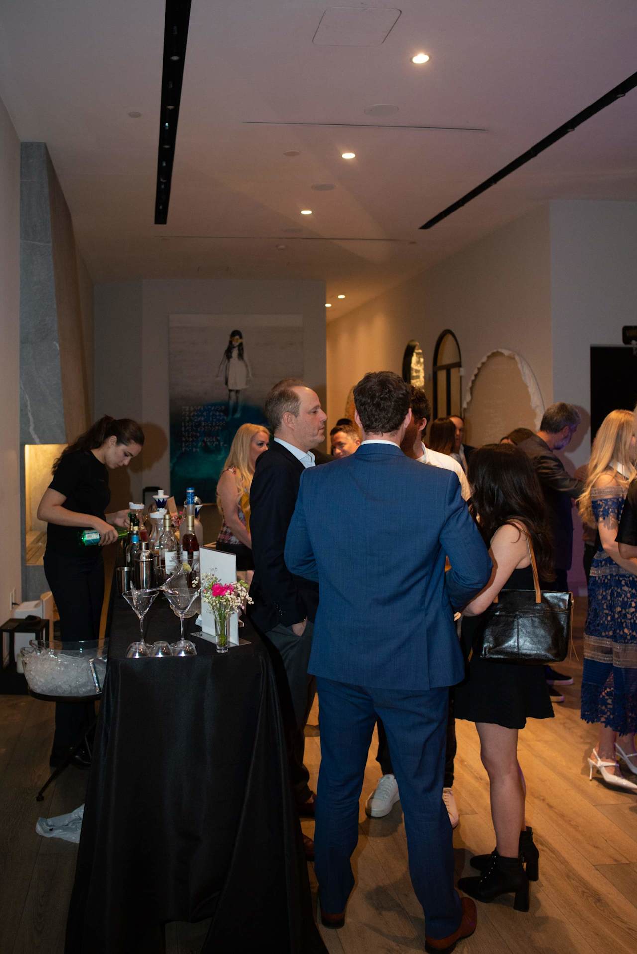 Guests mingle at an indoor event around a drinks table; a bartender pours a drink beside a man in a blue suit.