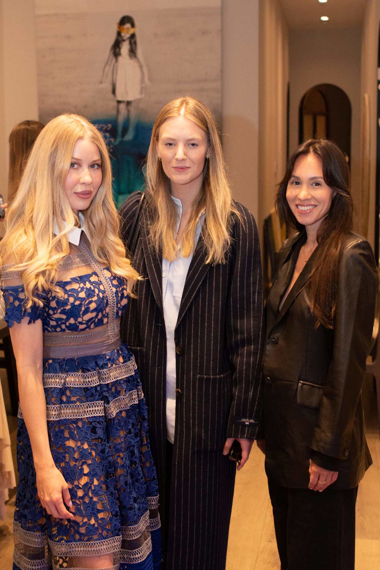 Three women pose together at an indoor event, wearing a blue lace dress, a pinstripe suit, and a black leather jacket.