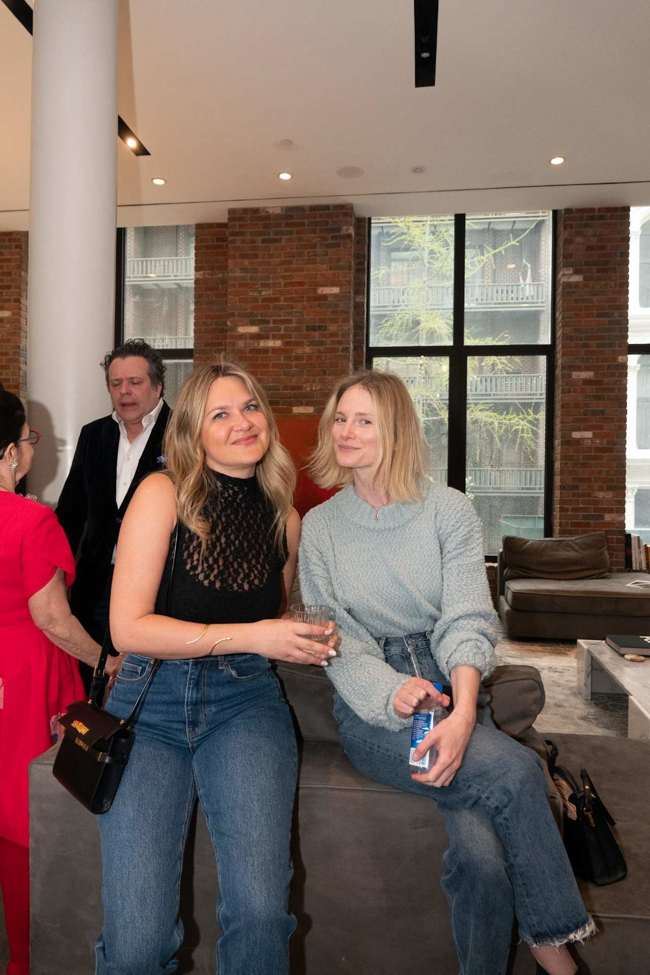 Two women sit together at a gathering in a brick-walled lounge, with other guests nearby.