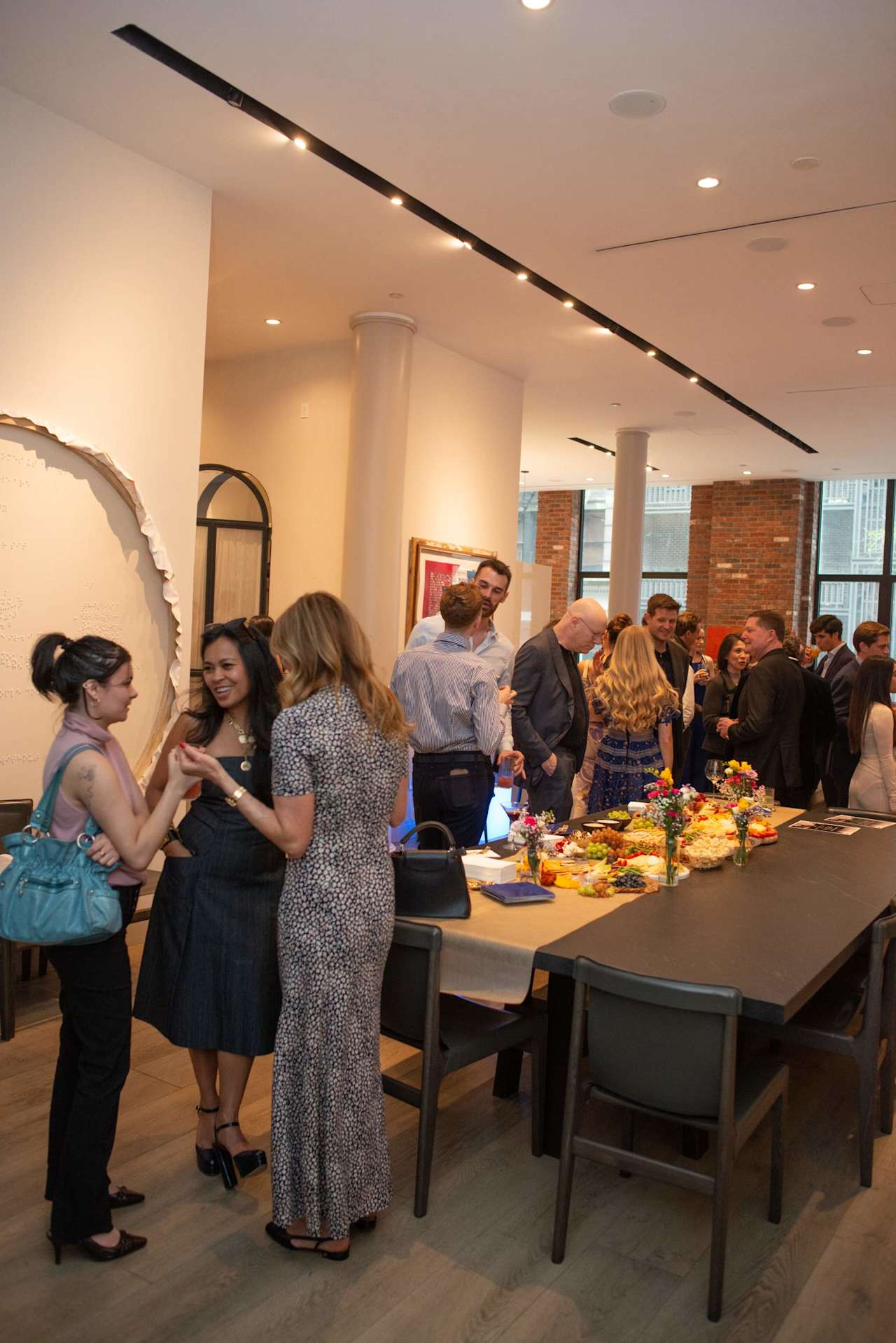 Guests mingle around a buffet table at a social gathering in a bright office with brick walls.