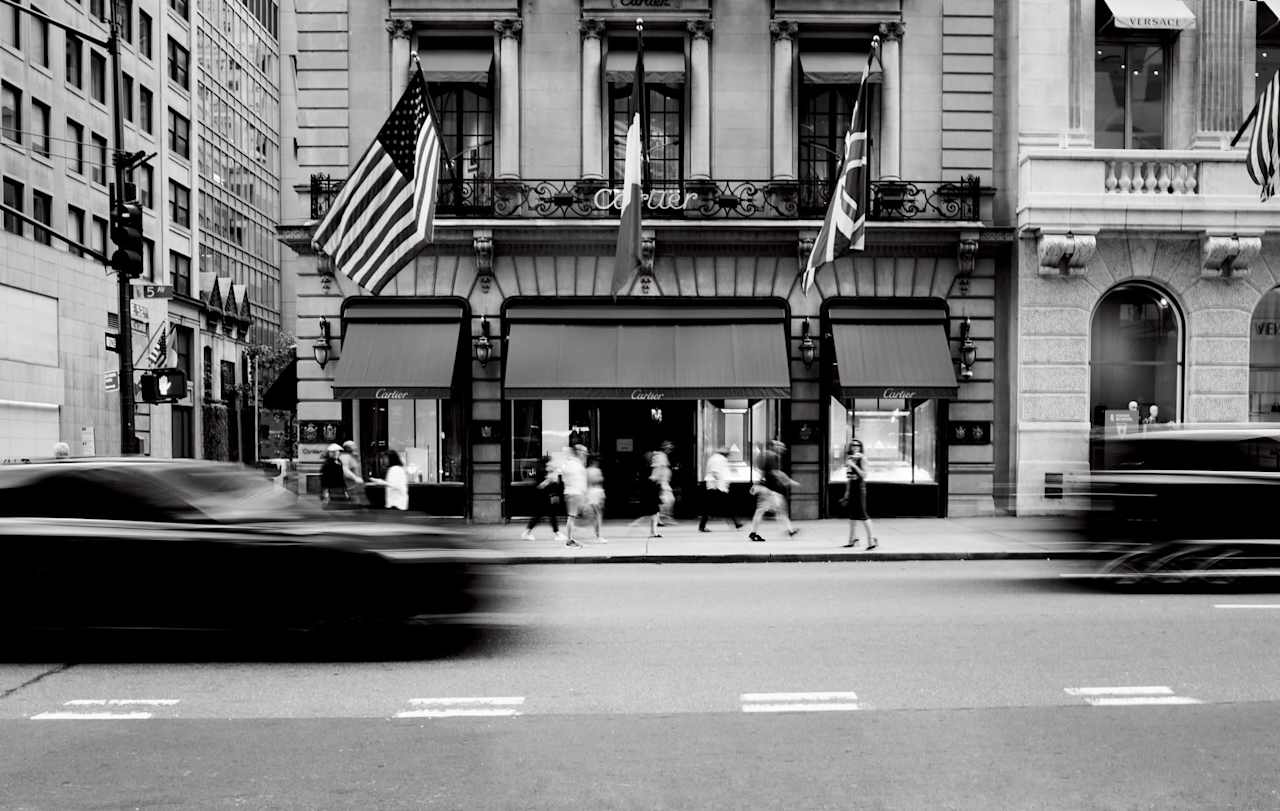 A black and white street scene featuring a Cartier store, with pedestrians walking and a car in motion. Flags are displayed above the entrance.