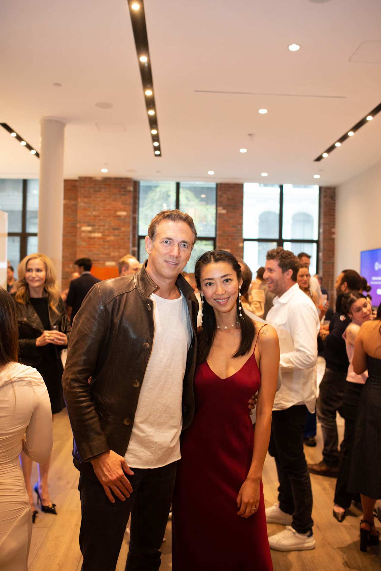 A man in a brown leather jacket and a woman in a red dress pose at a crowded indoor event.