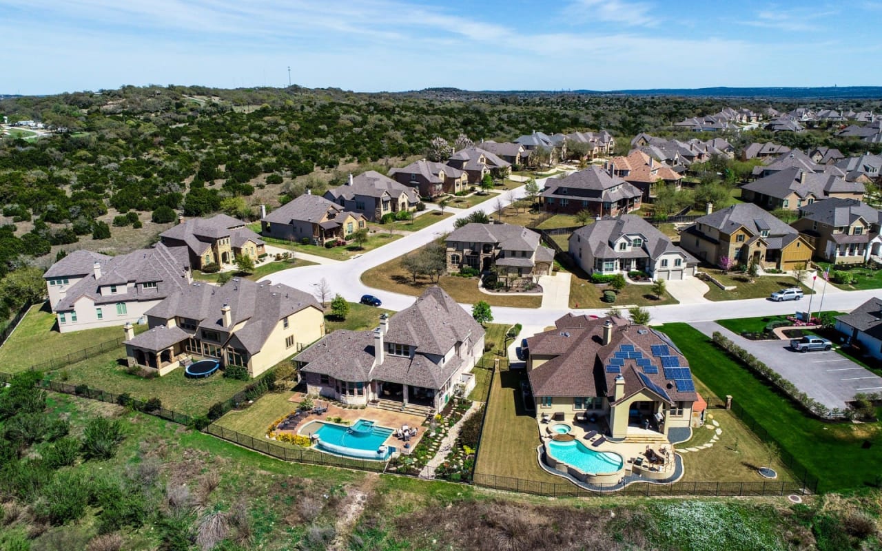 An aerial view of a residential area with houses, green lawns, driveways, garages, pools, and trees.