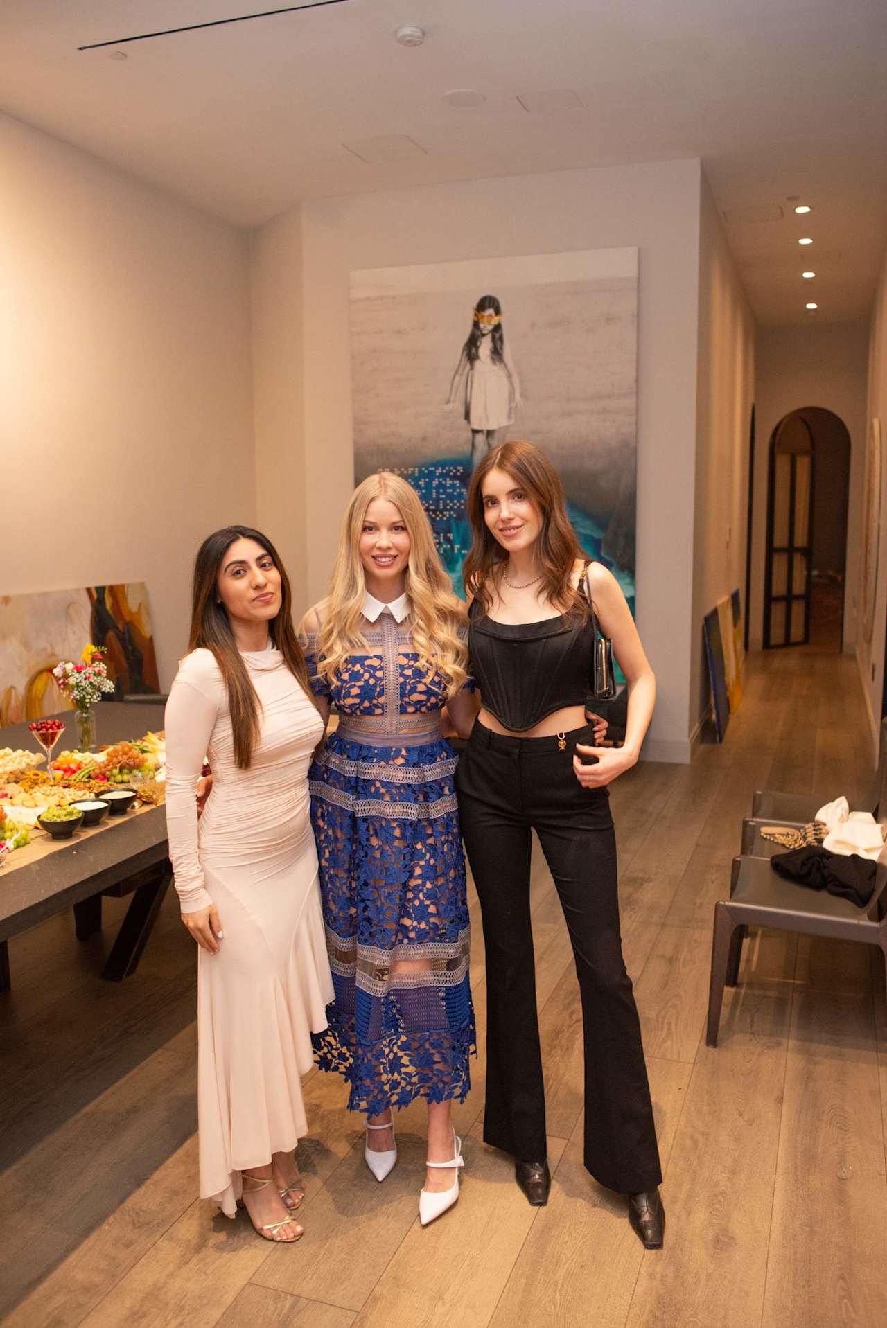 Three women pose together at an indoor event, wearing white, blue patterned, and black outfits.