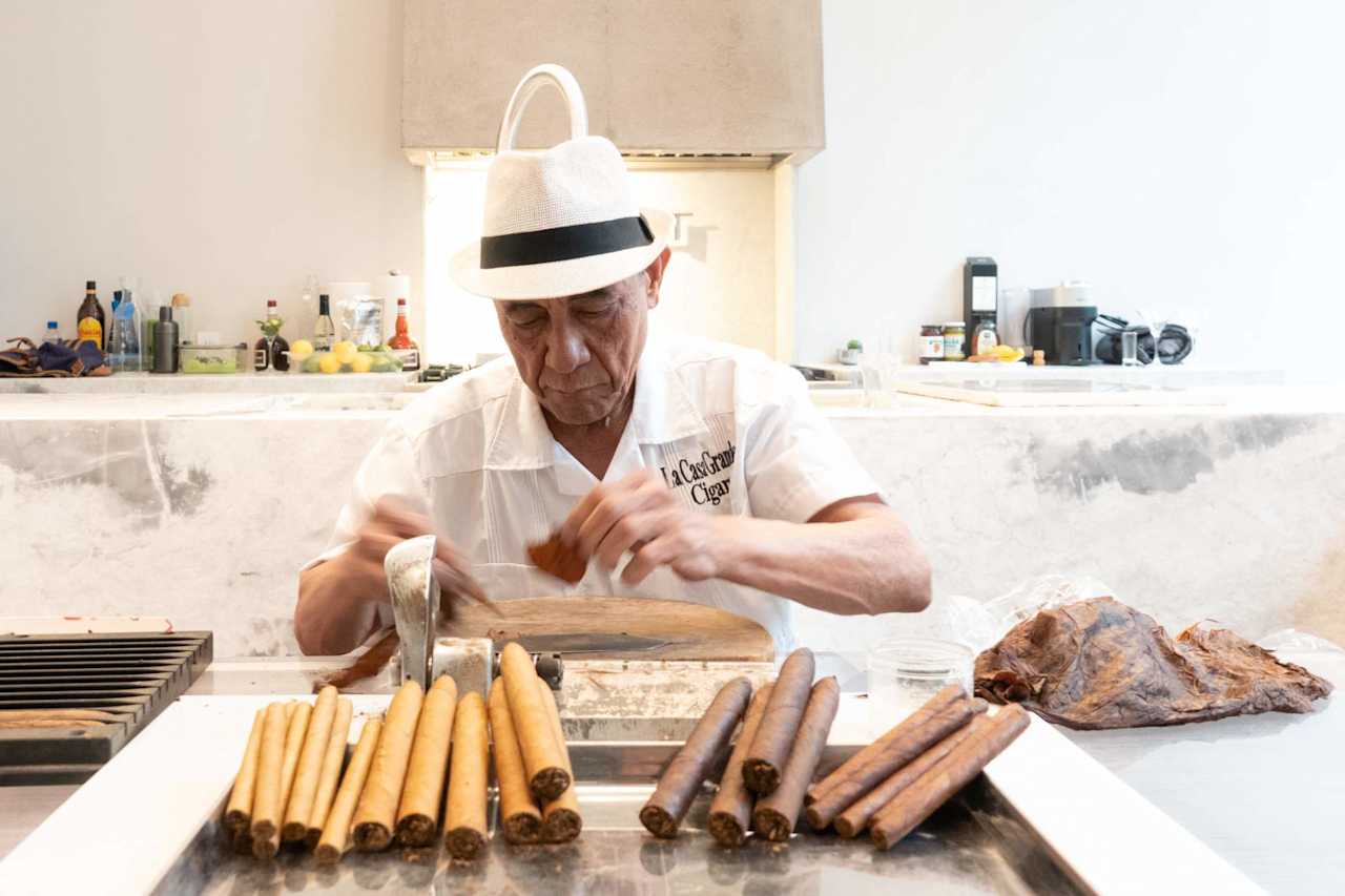 Cigar maker rolling cigars at a marble counter, with finished cigars arranged in front of him.