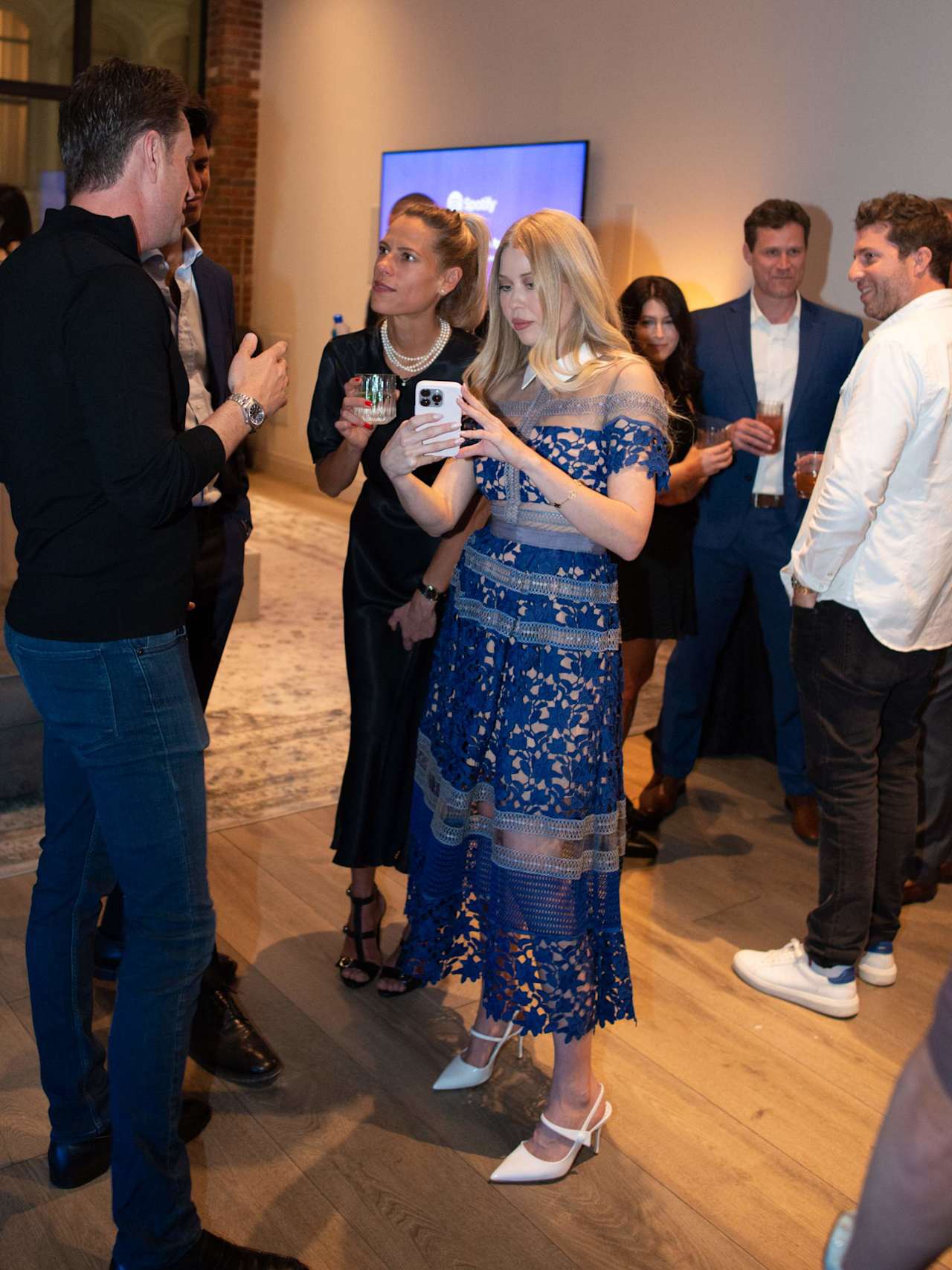 Guests mingle at an indoor event; a woman in a blue lace dress checks her phone.