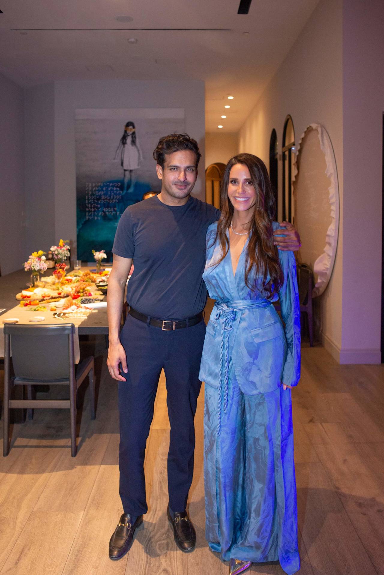 A man and woman pose together beside a buffet table; she wears a flowing blue patterned outfit.