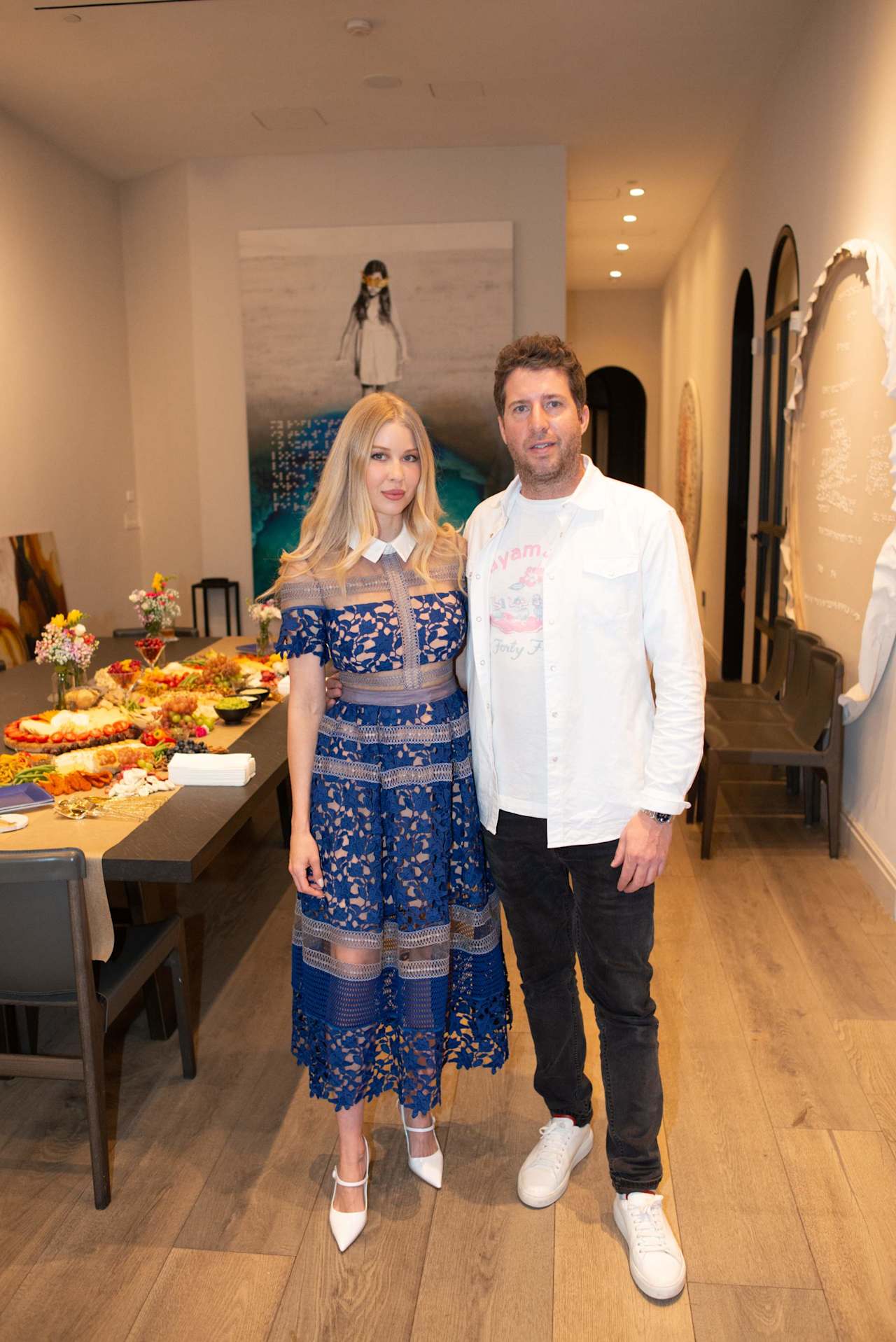A woman in a blue lace dress and a man in a white jacket pose beside a buffet table.