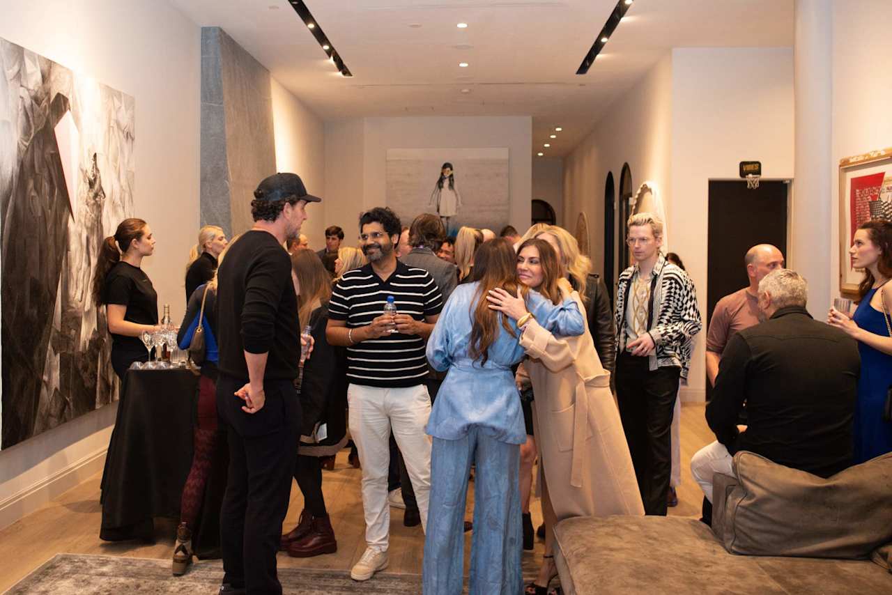 Guests mingle in an art gallery, including two women embracing near the center.