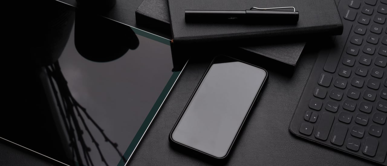 a black tabletop workspace with a tablet, a keyboard, a phone, and a notebook