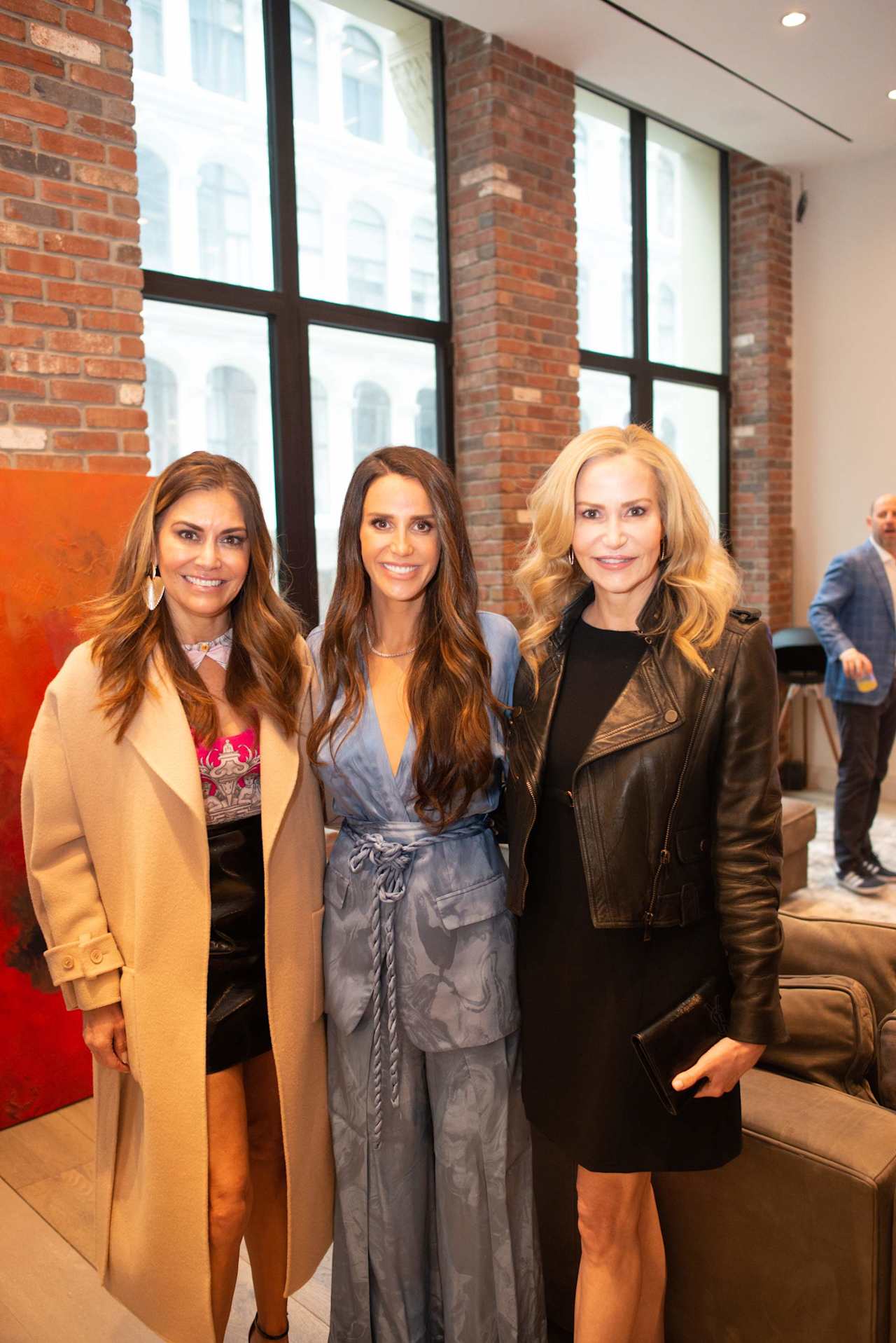 Three women pose together in a brick-walled office, with a man standing in the background.