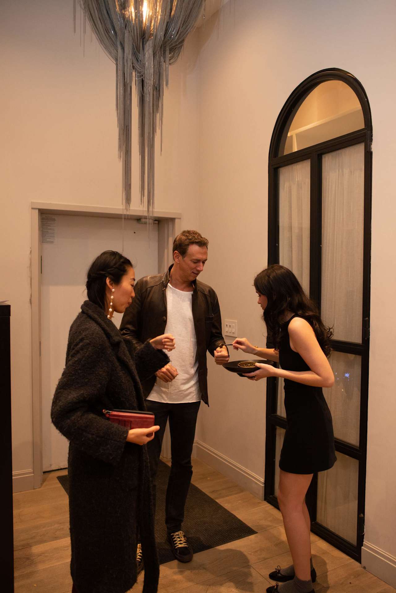 Guests mingle at an indoor gathering as a woman serves food beneath a sculptural chandelier.