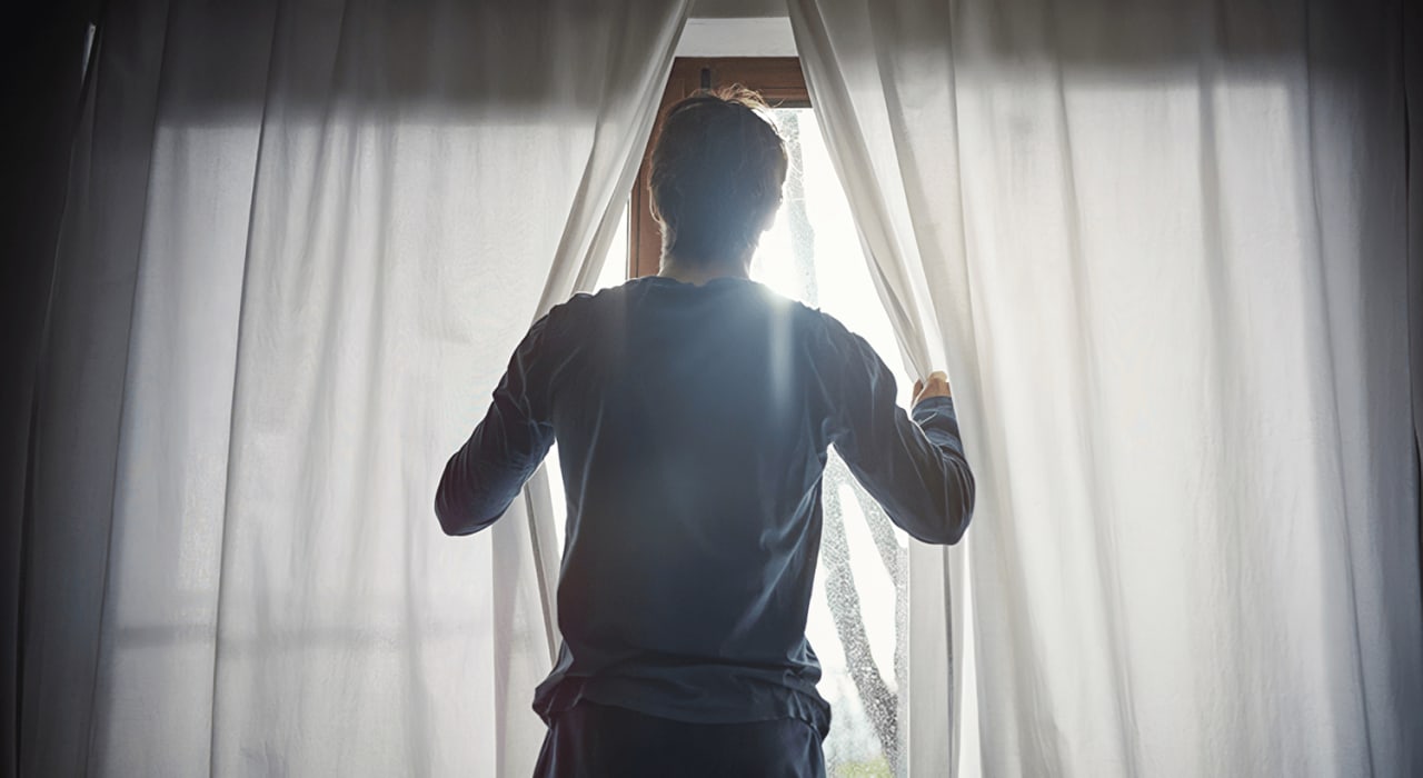 A guy in blue pajamas parting white curtains looking out the window