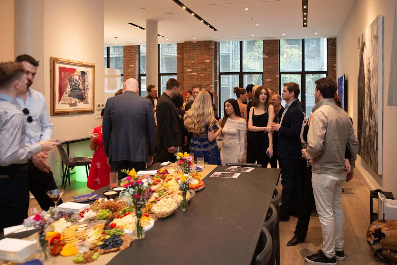 Guests mingle around a buffet at a real estate event in a brick-walled gallery.