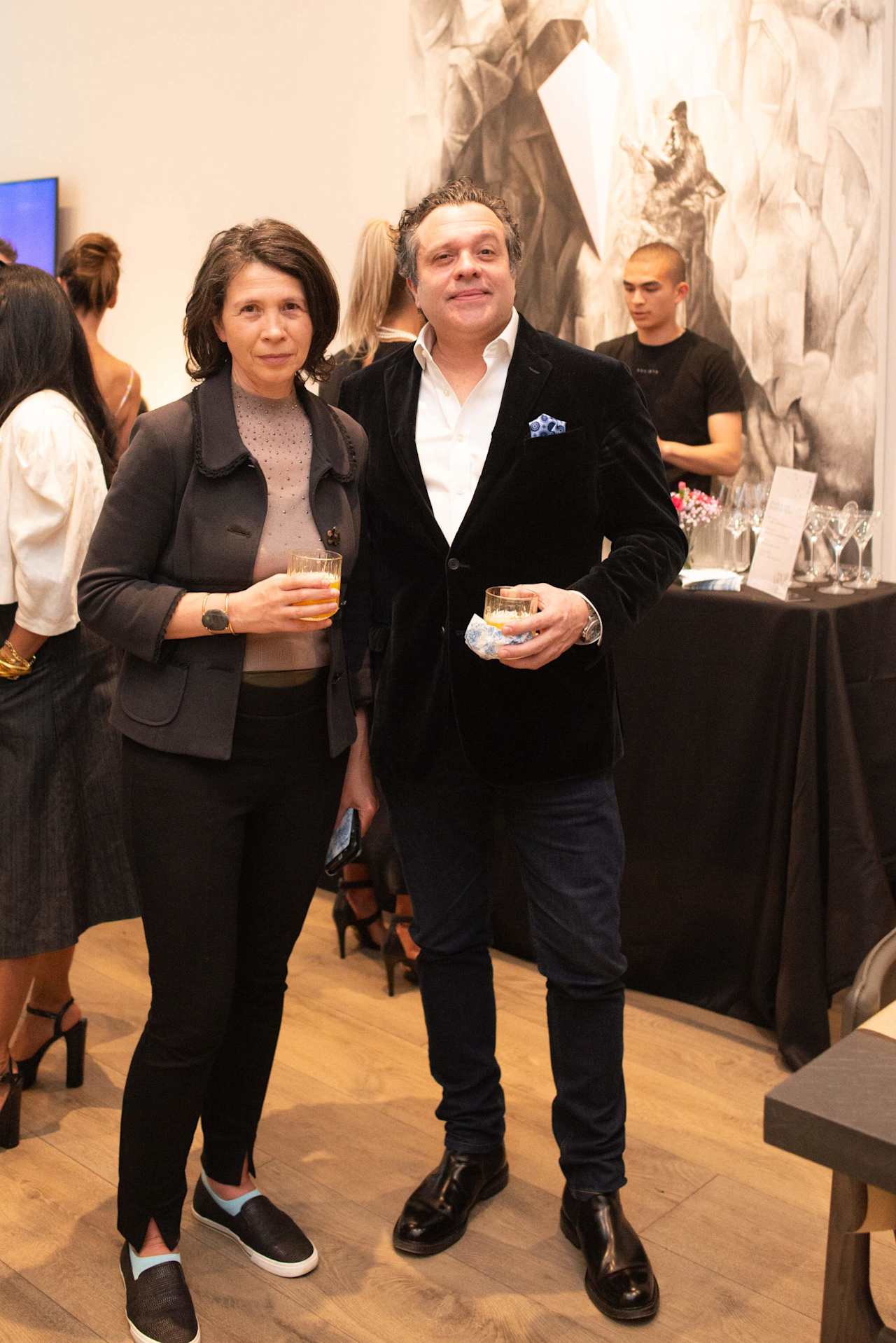 A woman and man pose with drinks at an event in front of an abstract mural.