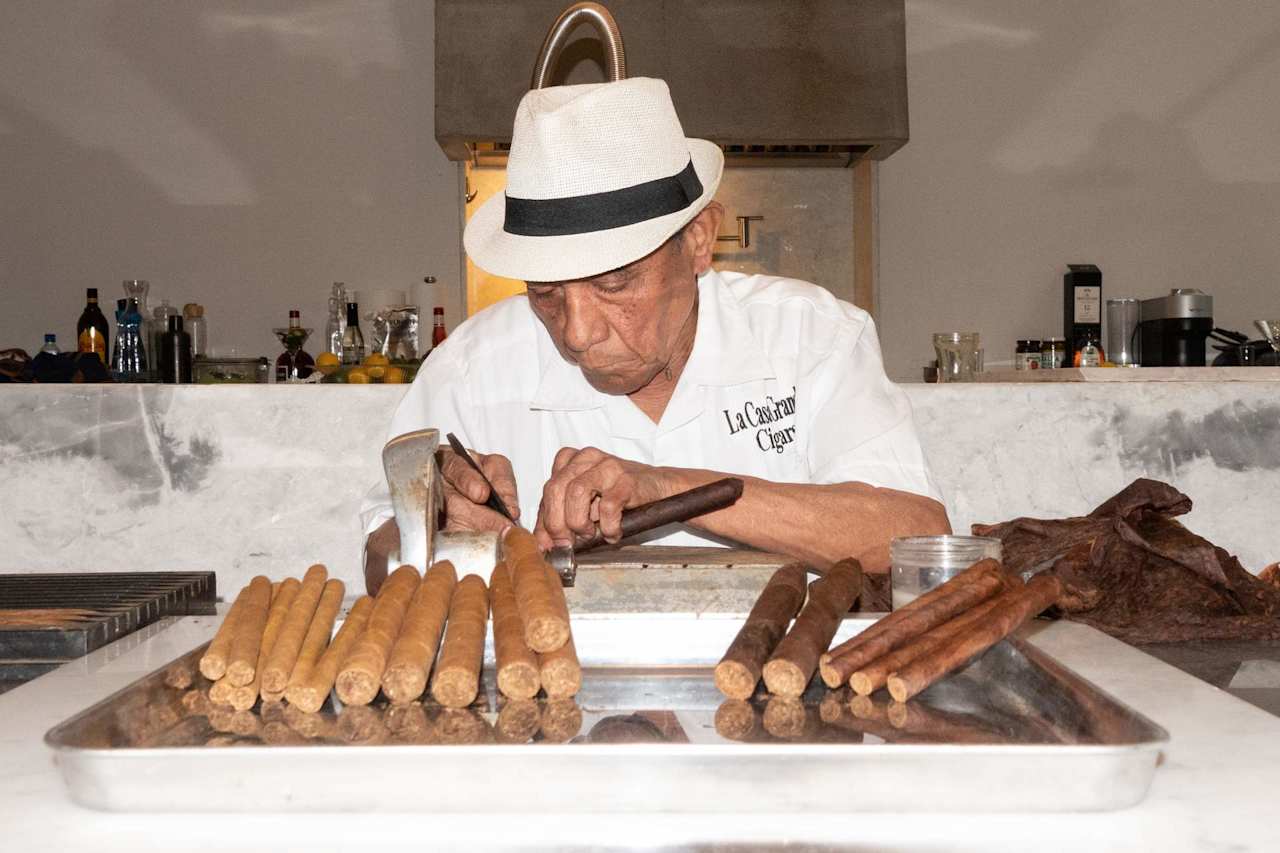 Man in a white fedora hand-rolling cigars behind a counter lined with cigars.