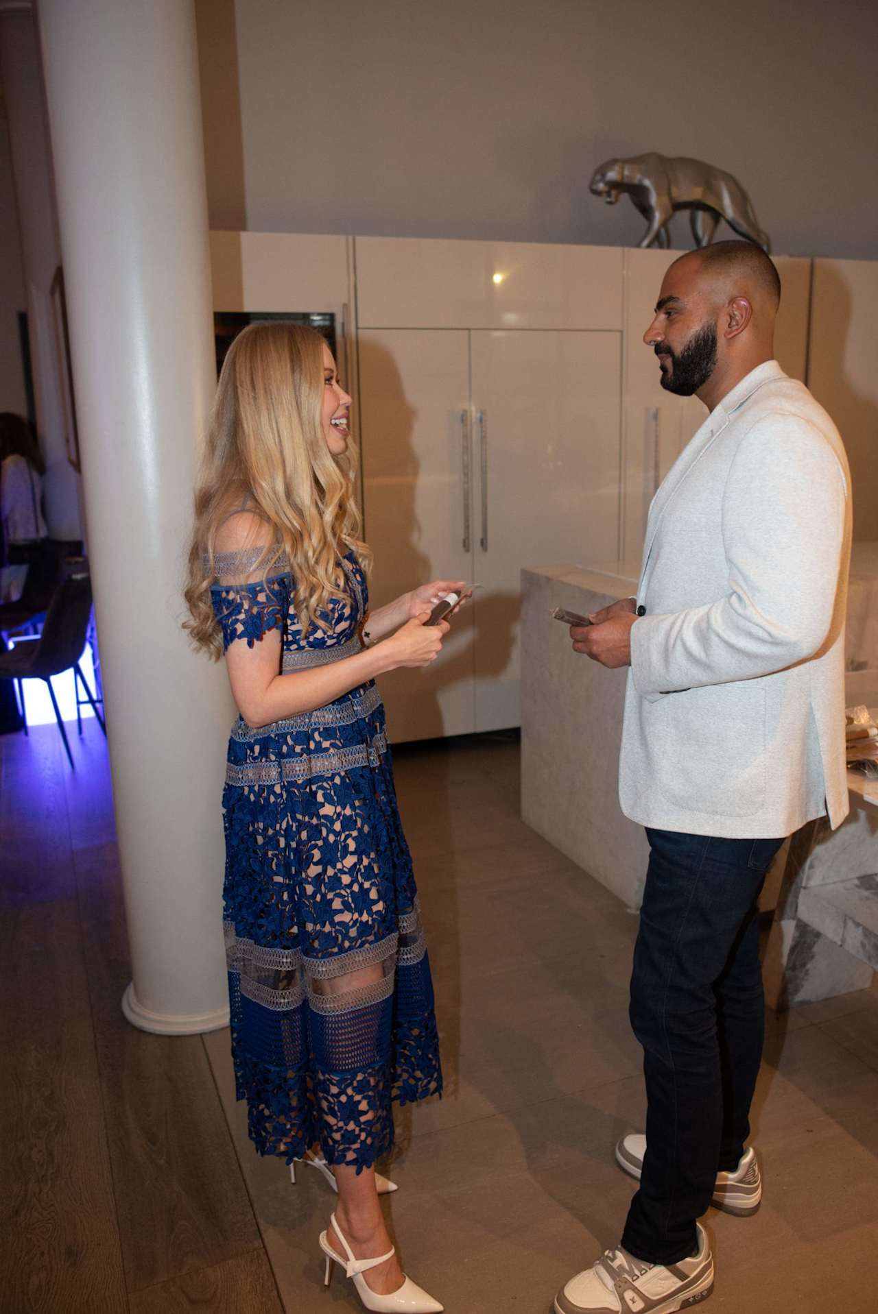 A woman in a blue lace dress talks with a man in a white jacket inside a modern room.