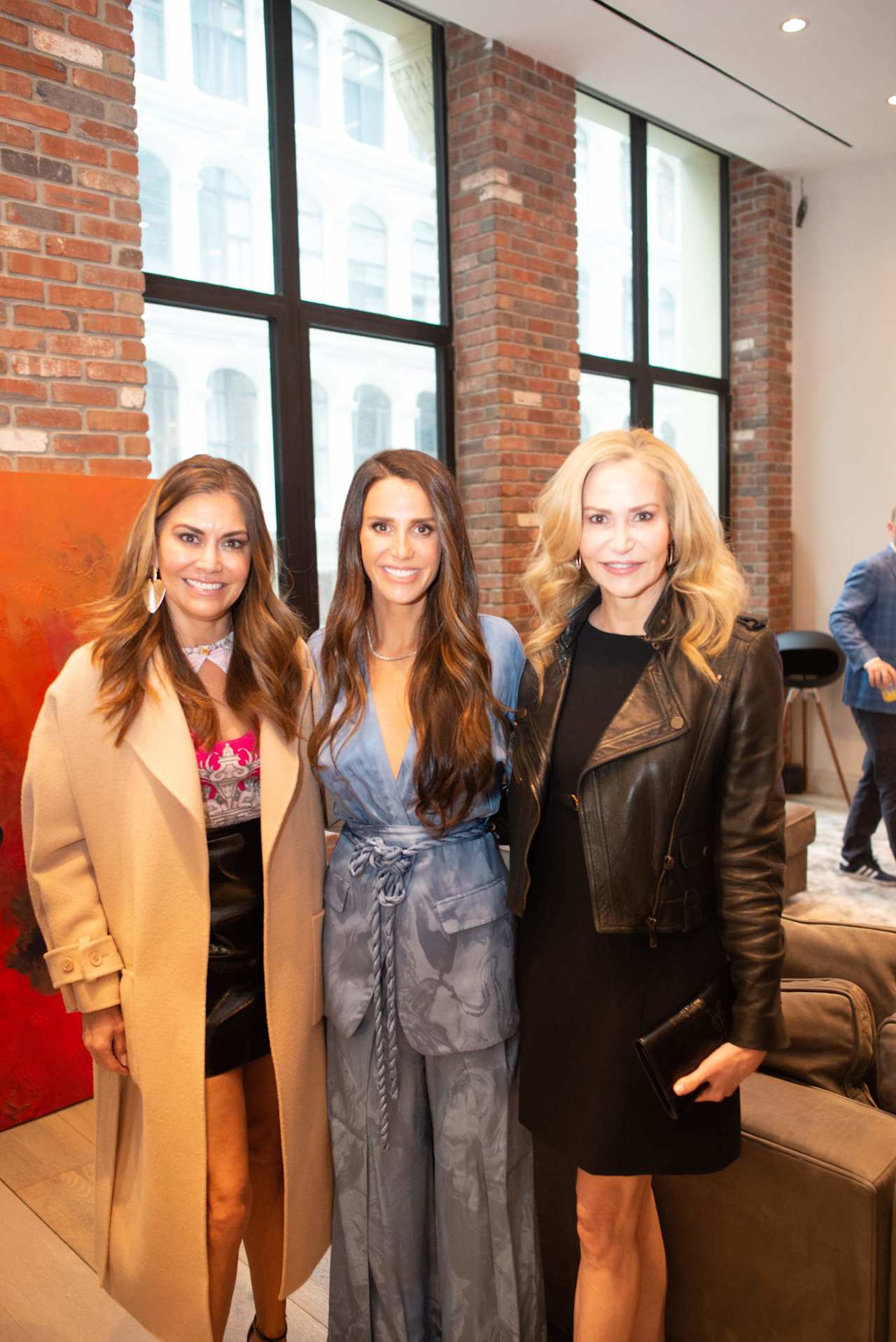 Three women pose together at an indoor event, with a partial view of another person at the right edge.