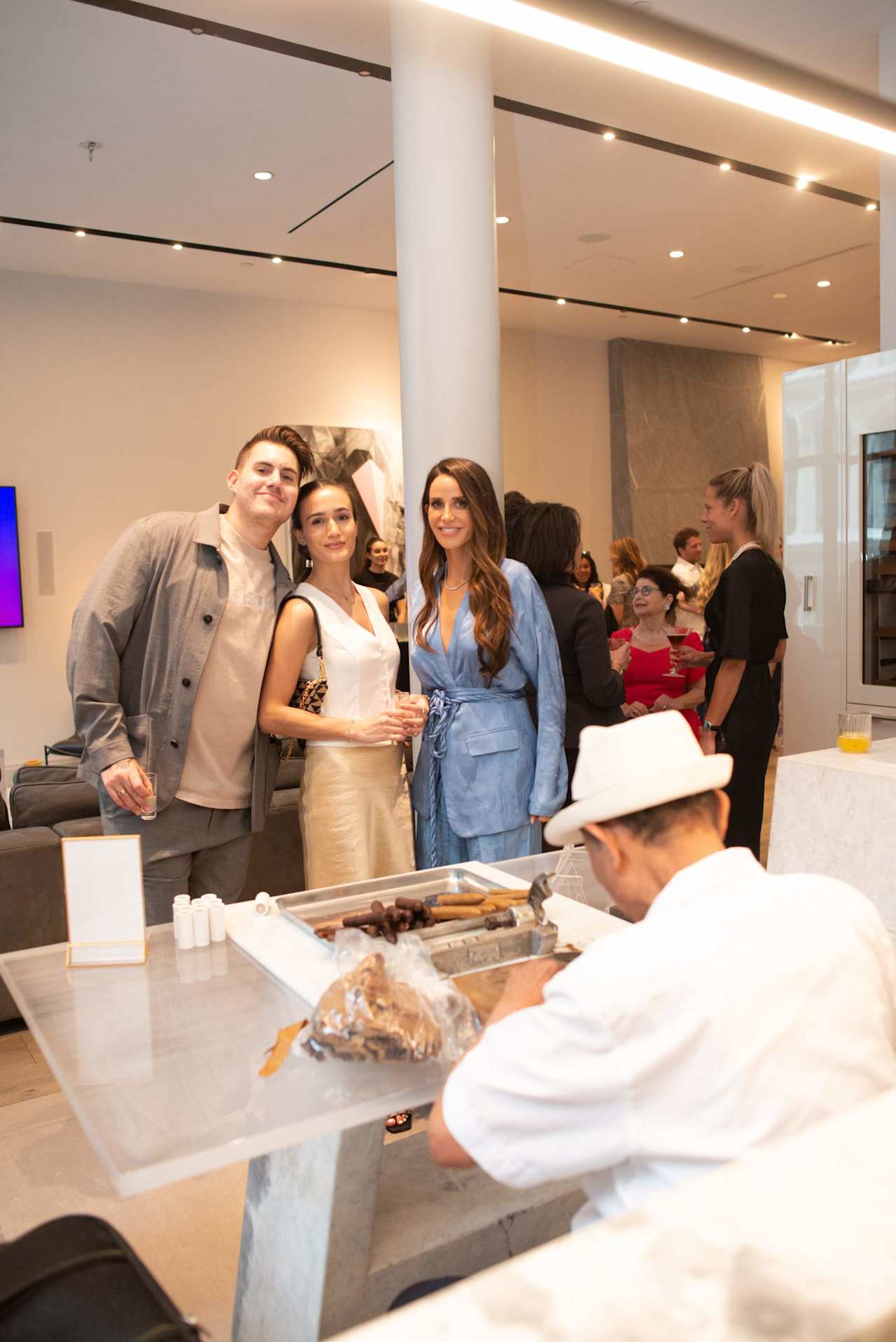 Guests at an indoor event gather around a food table as three attendees pose together.