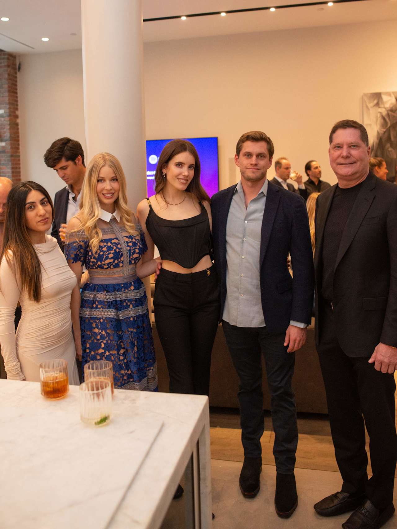 Six guests pose together at a reception, with other attendees and drinks on a marble counter.