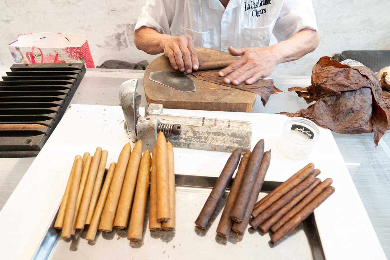 A cigar maker rolls tobacco leaves beside rows of finished cigars.