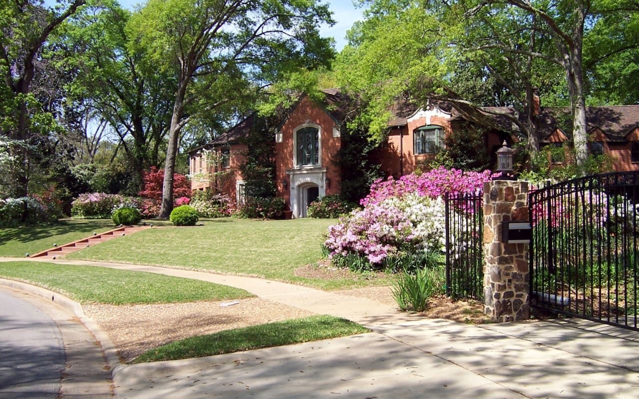 A single-family brick house with a front lawn, flower beds, a paved walkway from the sidewalk, and a gated entrance.