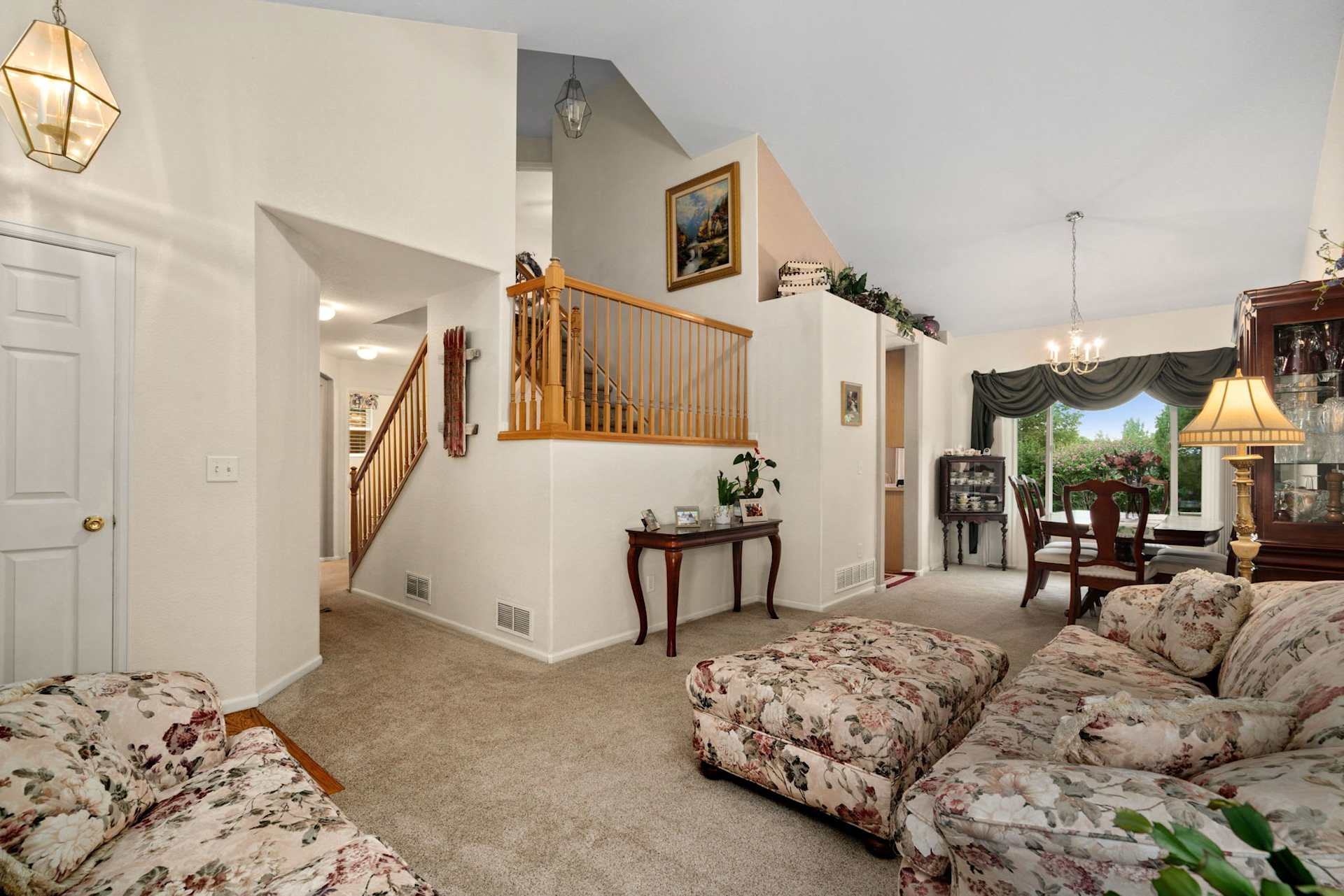 Living room with floral sofas, a wooden staircase, and dining area beside large windows.