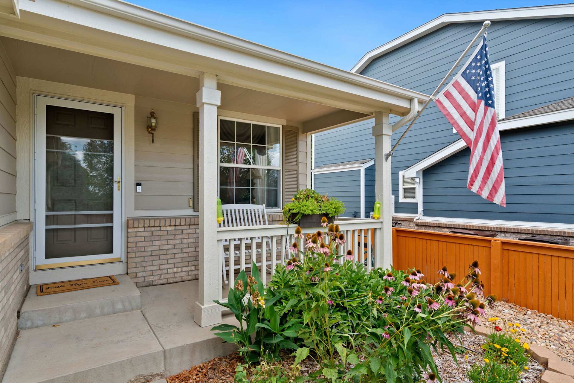 Covered home entrance with an American flag, porch bench, and flowering garden.