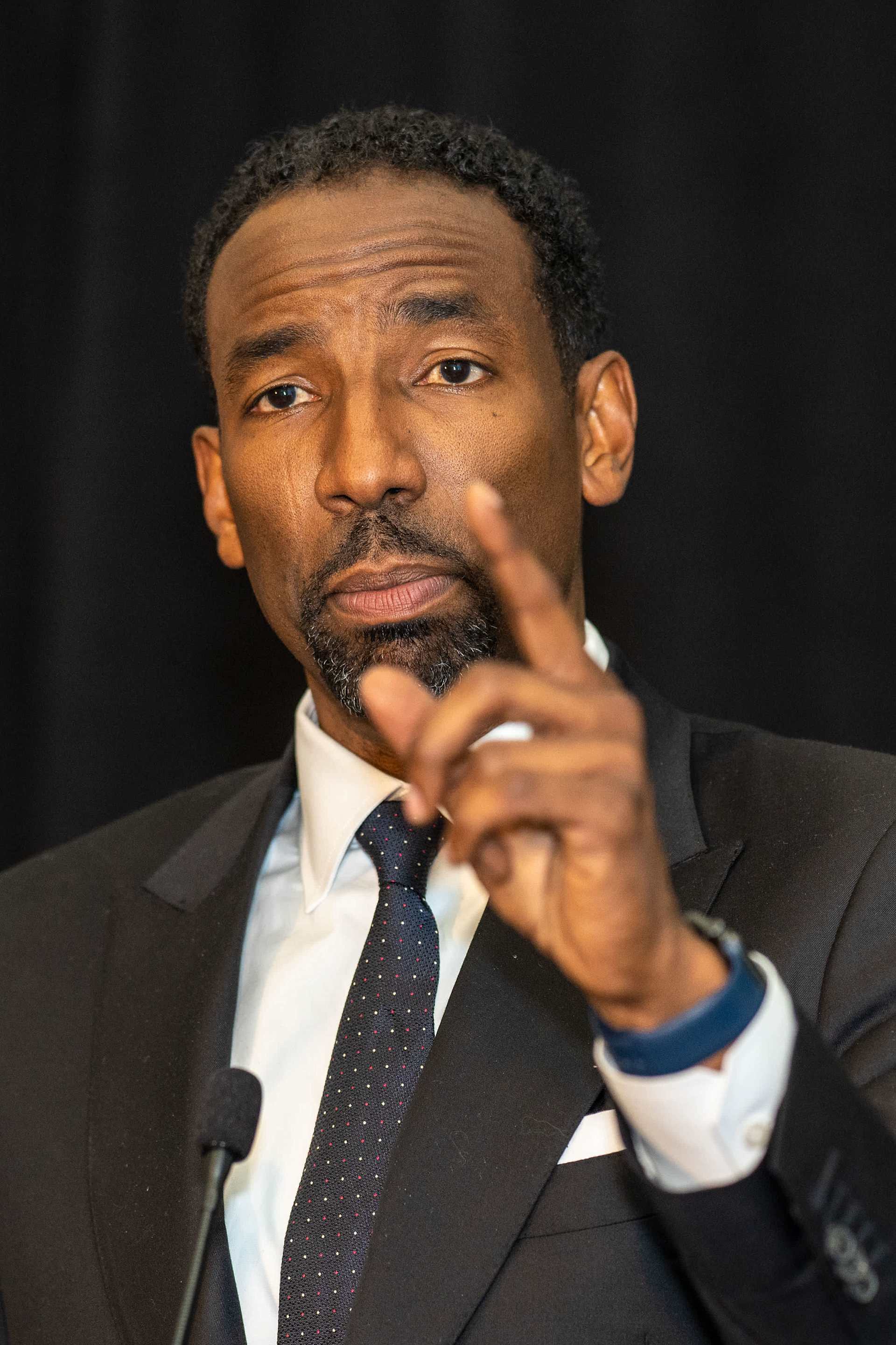 A man in a suit gestures with one hand while speaking, set against a dark background.