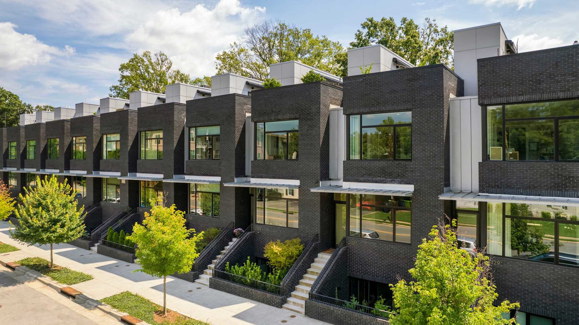 A row of modern black brick townhouses with large windows and greenery in front, under a blue sky with scattered clouds.