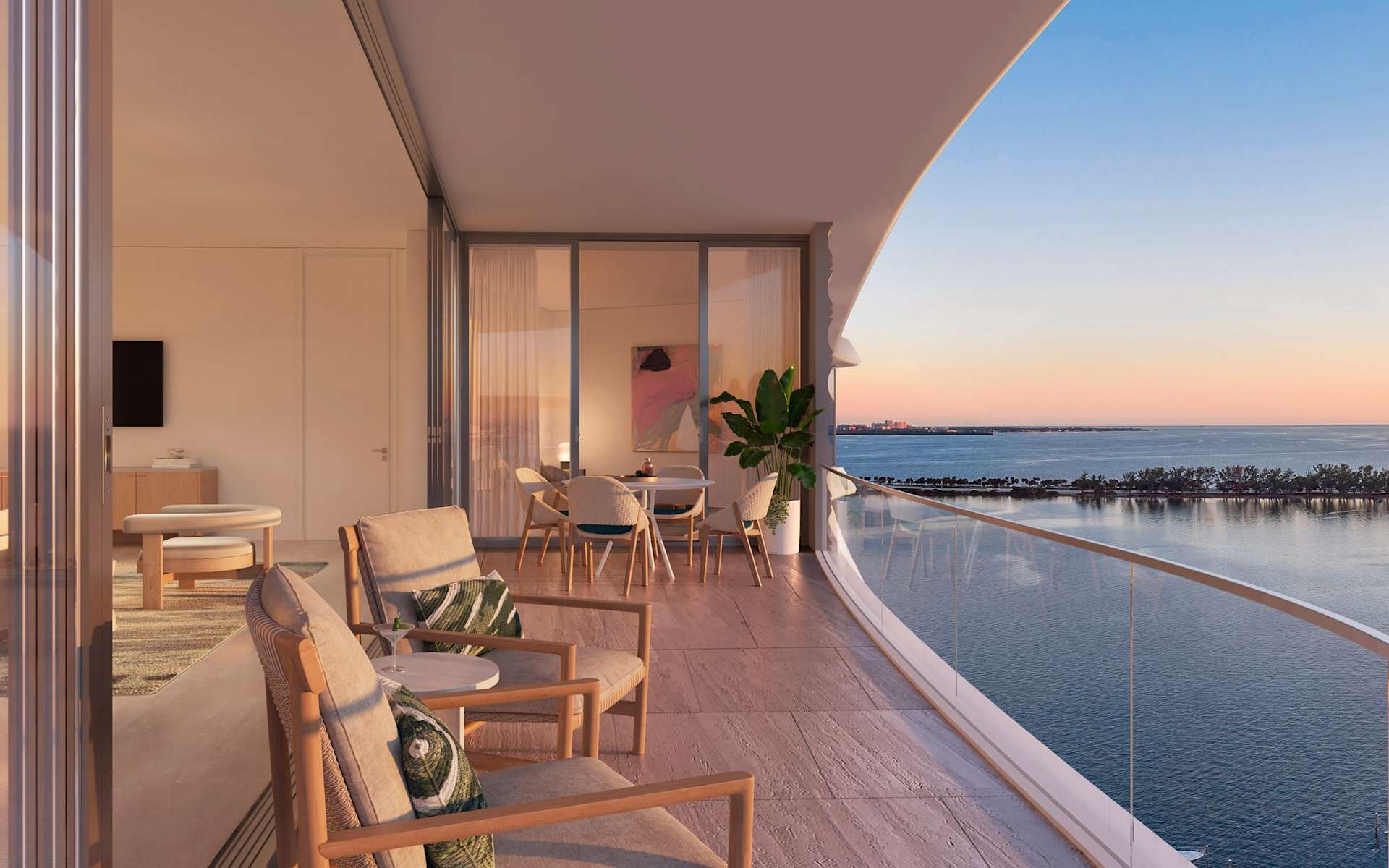 Furnished residence balcony overlooking water at sunset, with a curved glass railing and outdoor dining area.